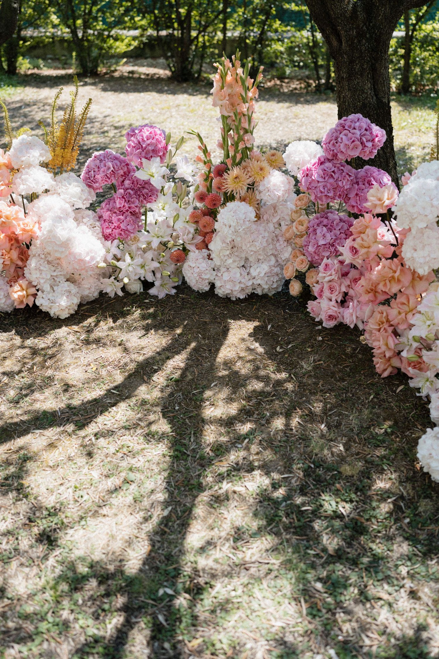 Lush pink and white floral arrangement with hydrangeas and orchids displayed outdoors under a tree at a wedding.