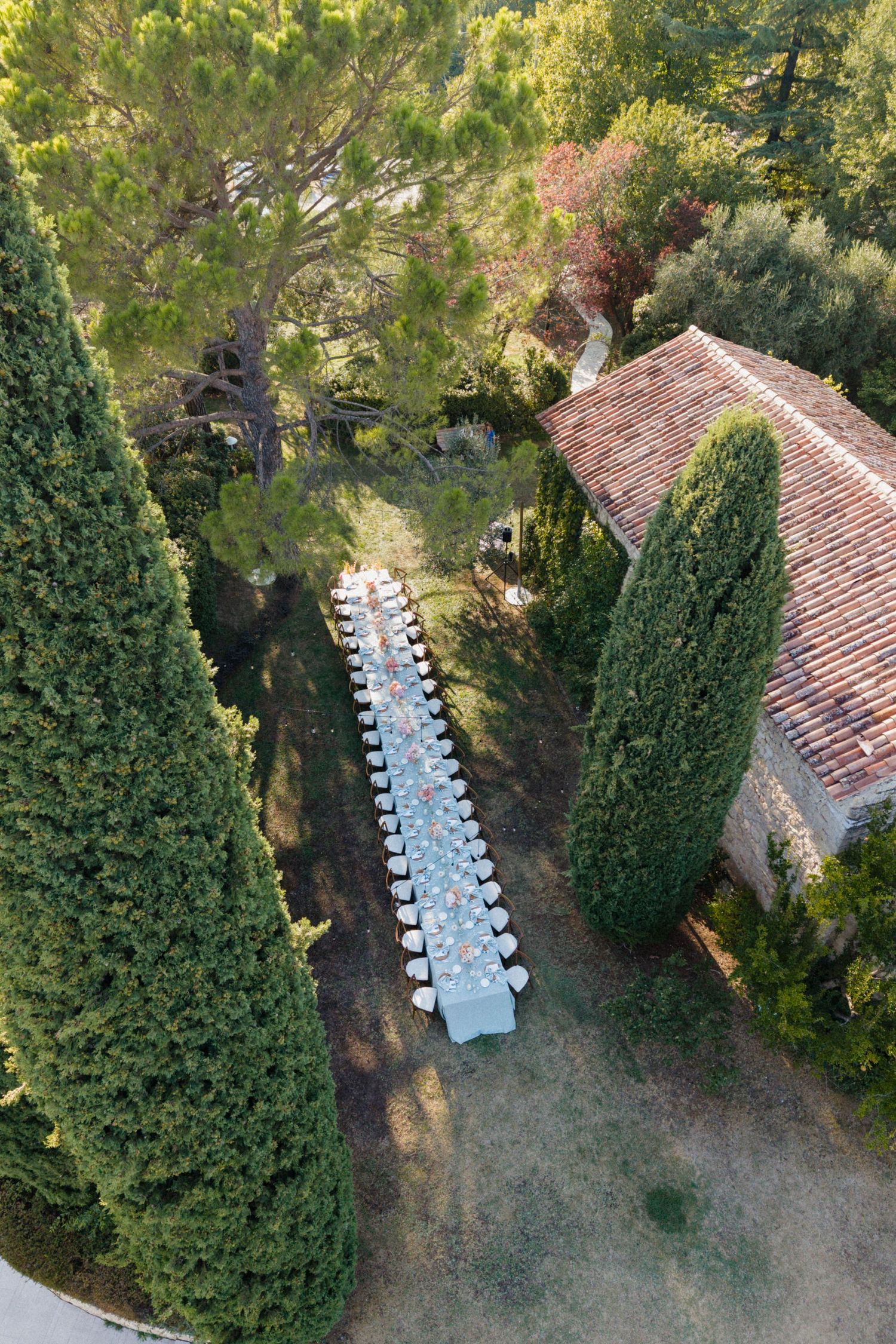 Aerial view of a long outdoor dining table set for an event, surrounded by cypress trees and a stone building.