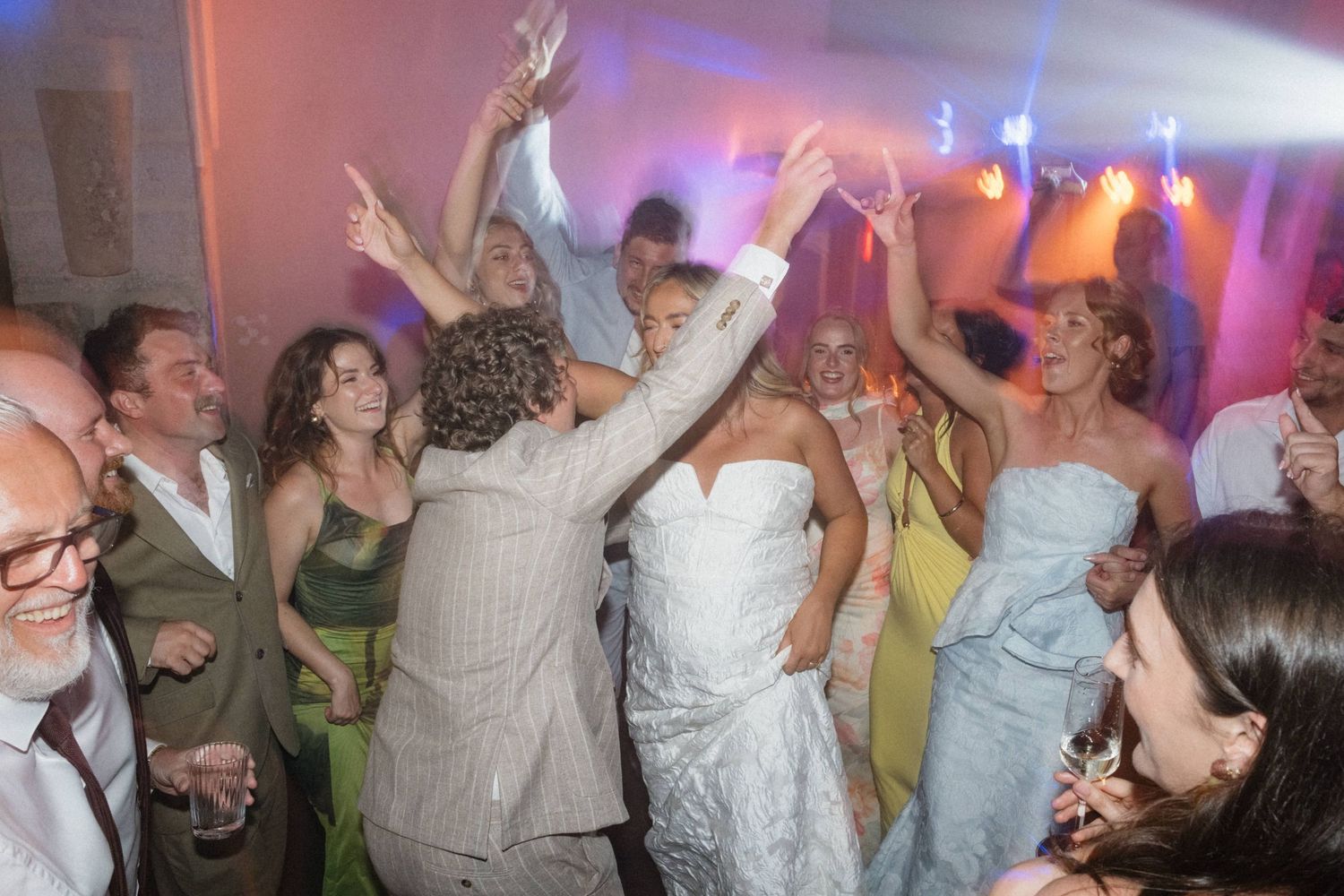 Bride and groom dancing joyfully with wedding guests under colorful lights at a lively wedding reception.