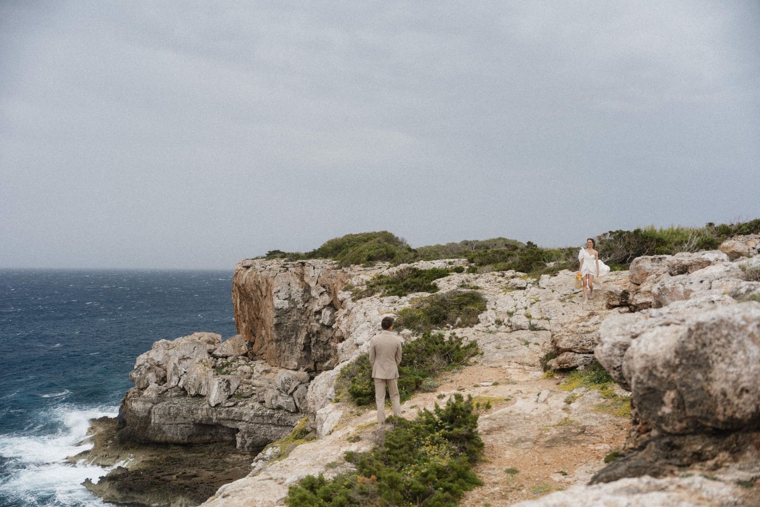Two people stand on rugged coastal cliffs overlooking a turbulent ocean under an overcast sky.