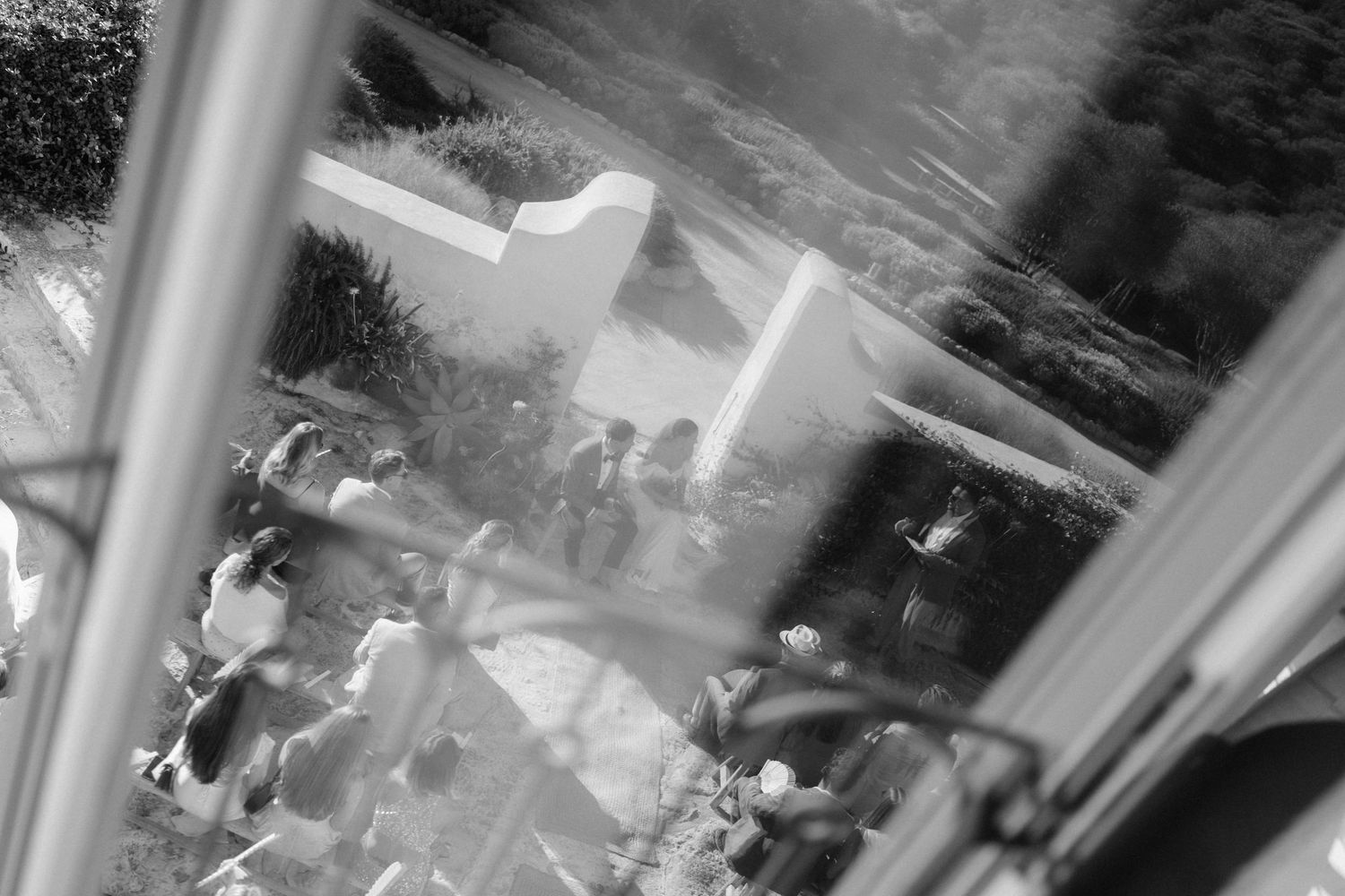 Black and white reflection in a window shows wedding guests gathered outdoors near a curved white wall.