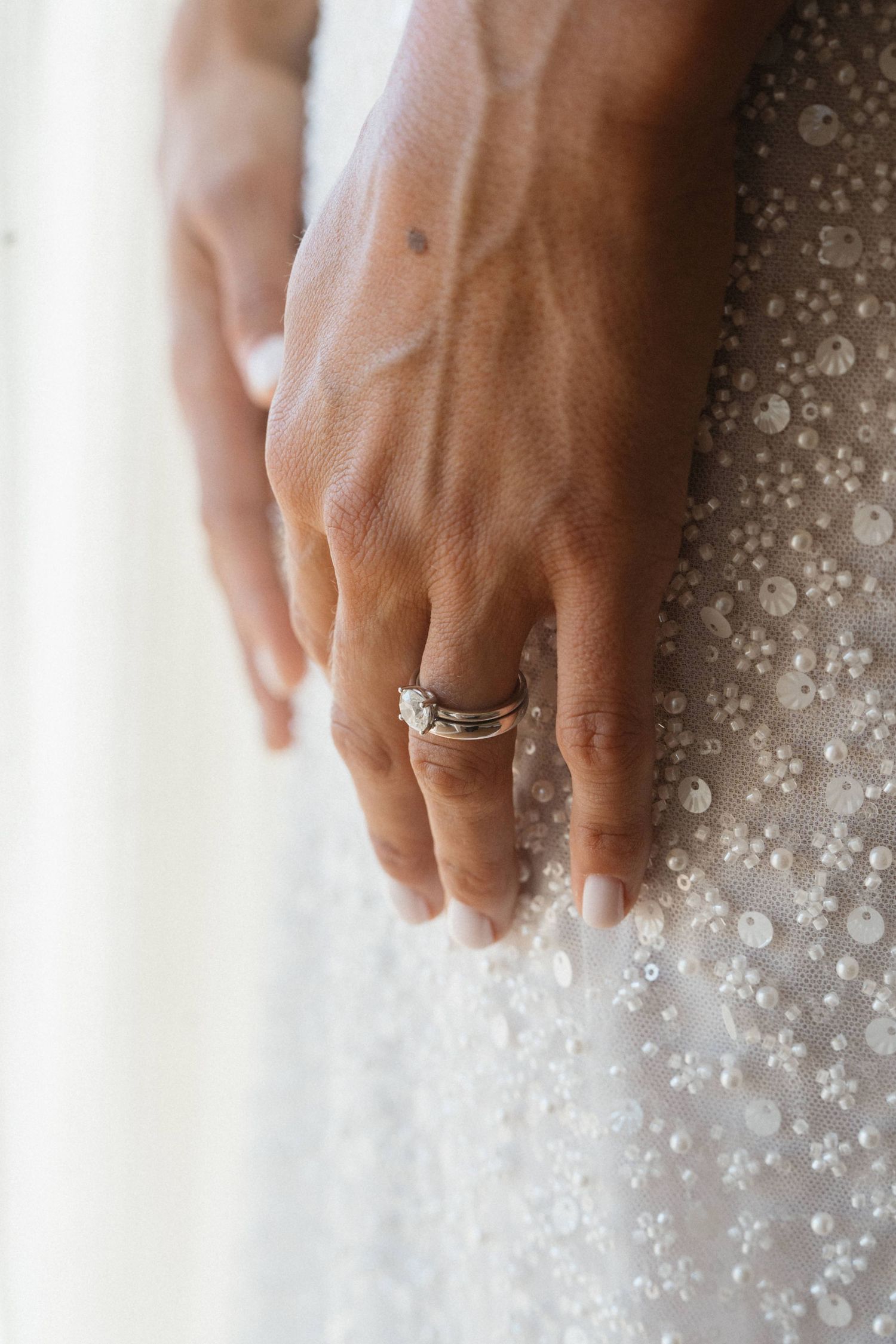 Close-up of a bride's hand with a diamond engagement ring and wedding band resting on a beaded white wedding gown.