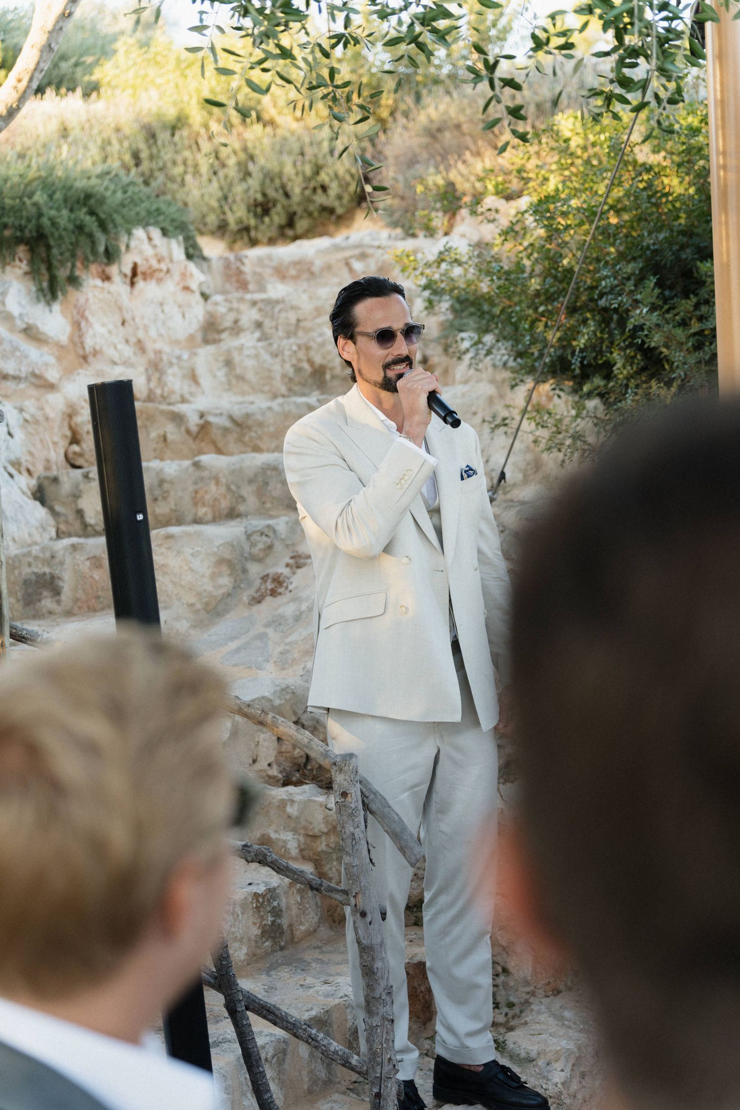 A man in a cream suit and sunglasses speaks into a microphone at an outdoor wedding ceremony beside a rustic stone wall.