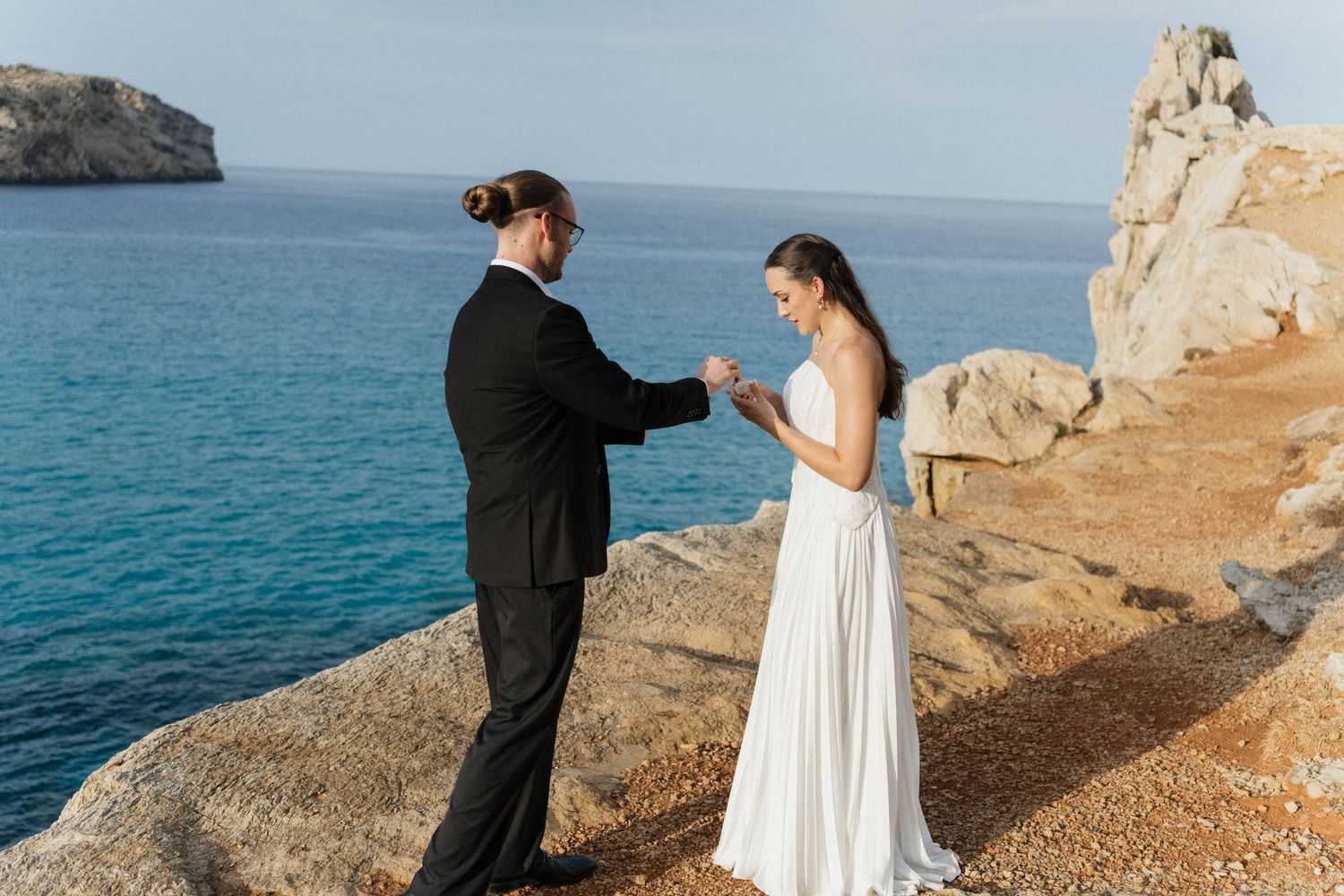 Couple exchanging vows on a rocky coastal cliff above the turquoise sea, the bride in a flowing white dress and the groom in a black suit.