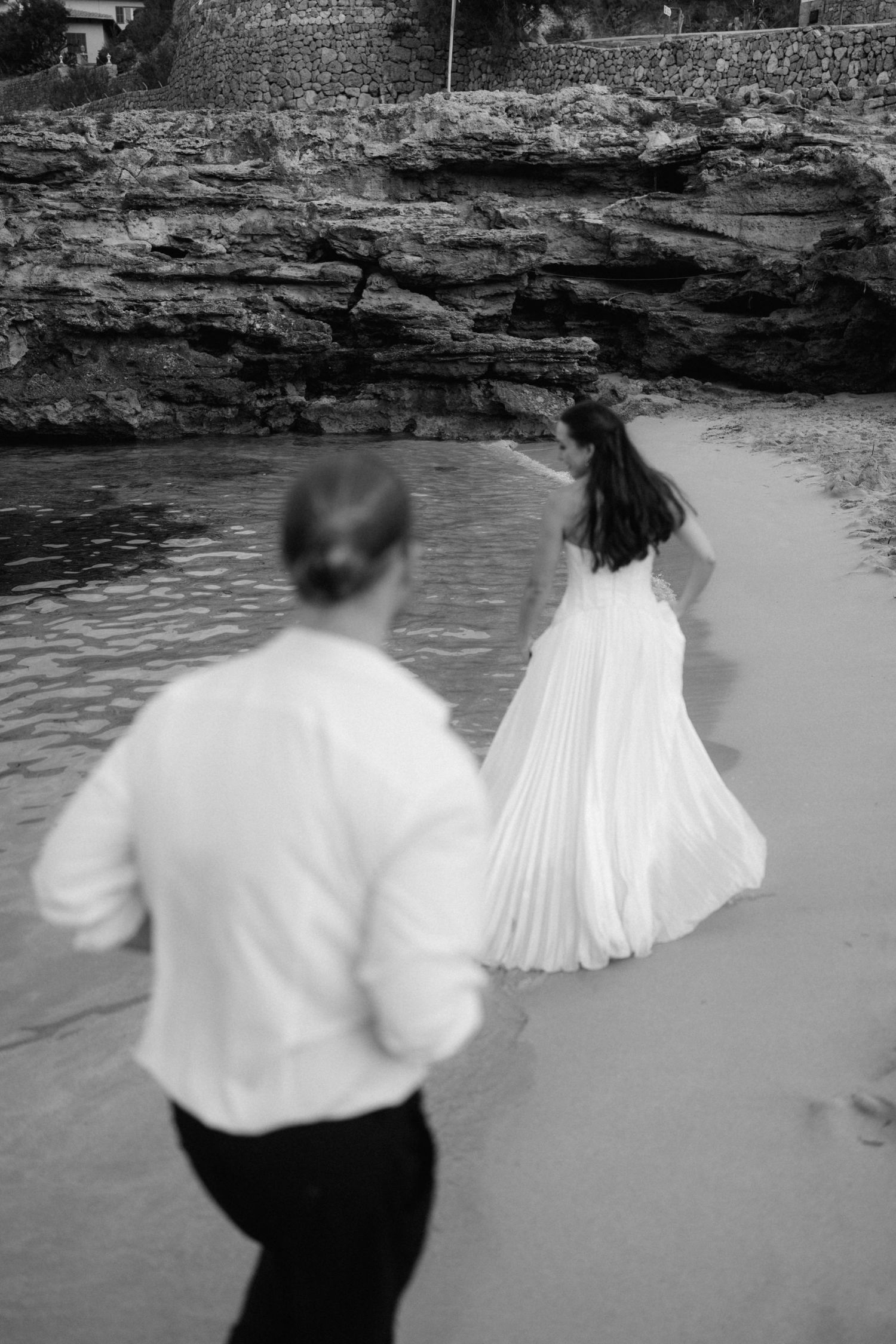 Black and white photo of a bride in a flowing white gown walking along a rocky beach shoreline as a blurred groom follows.