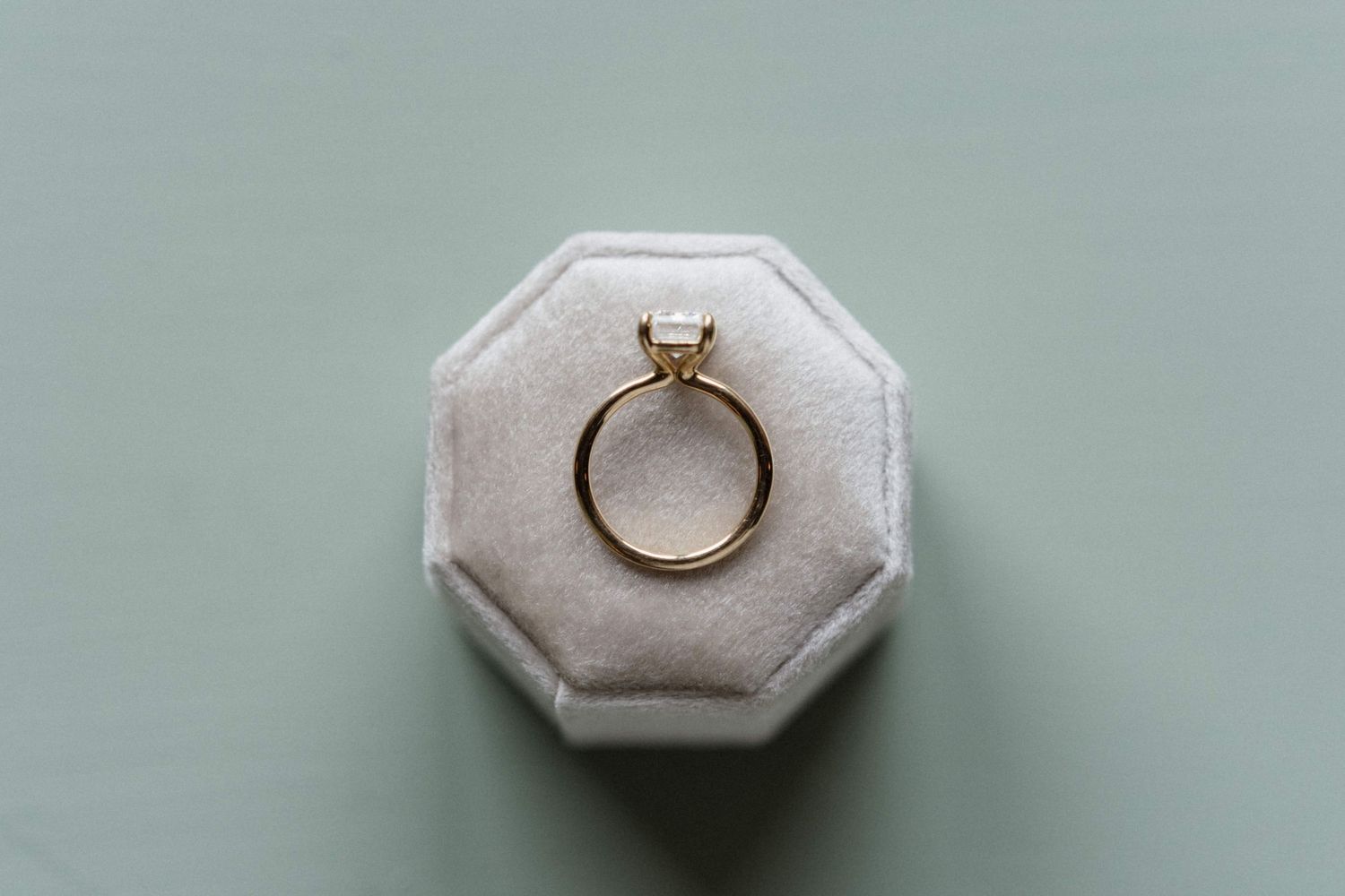 Gold solitaire engagement ring with an emerald-cut diamond resting in a gray octagonal velvet ring box on a soft sage green background.