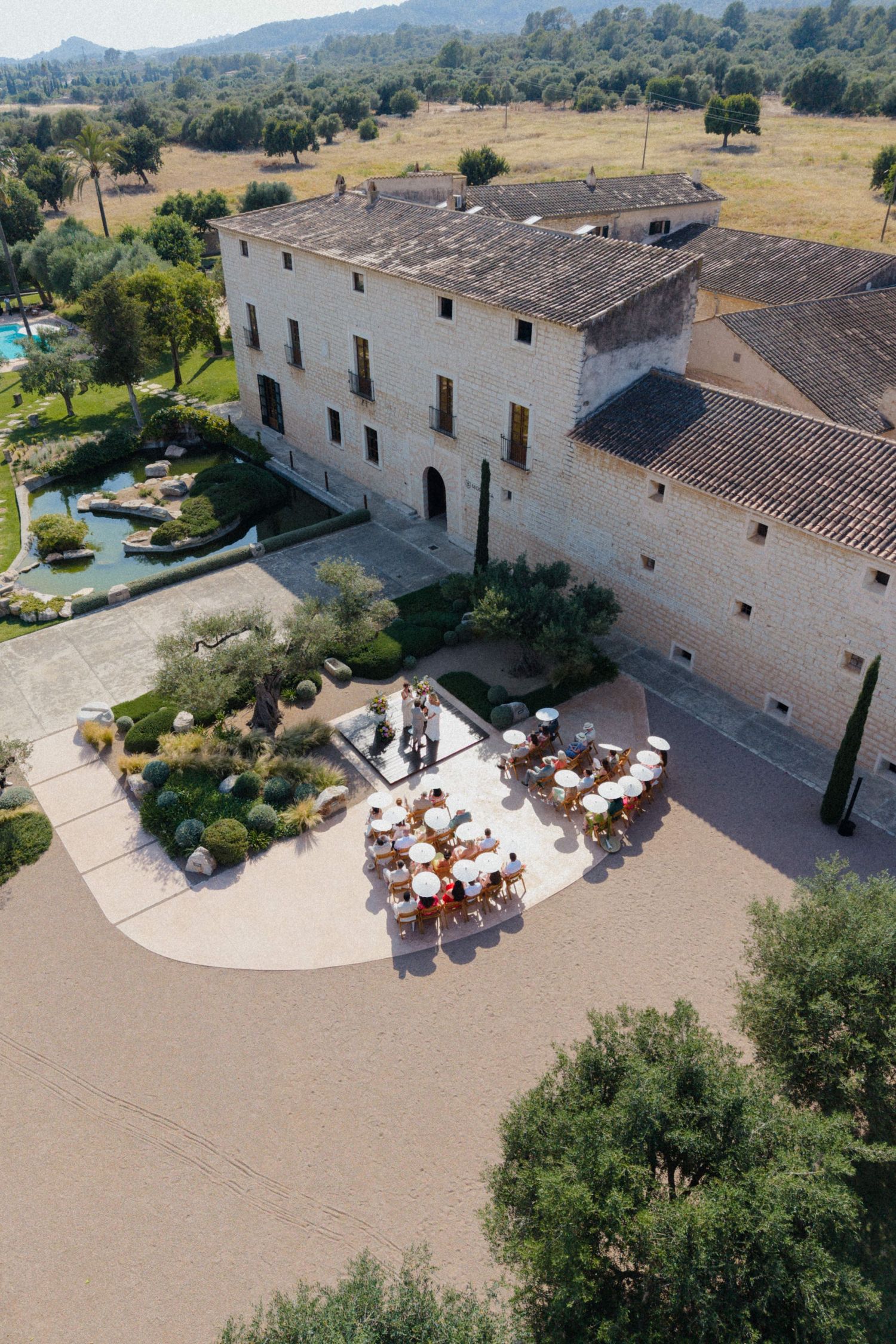 Aerial view of a rustic stone villa with outdoor dining terrace, lush gardens, and pool in a Mediterranean landscape.