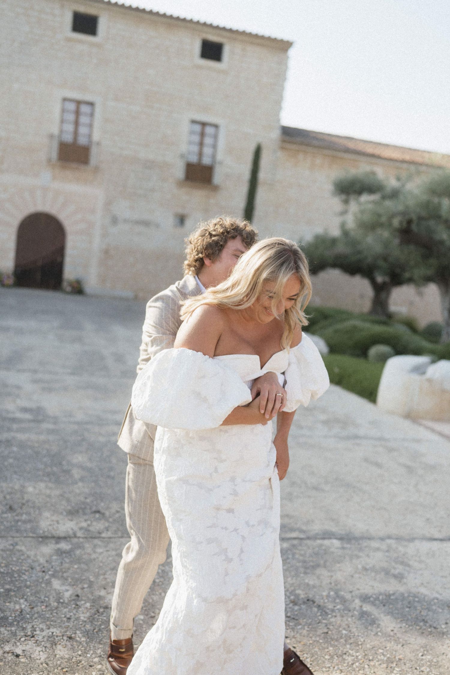 Bride in off-shoulder white gown embraced by groom in linen suit outside a stone villa.