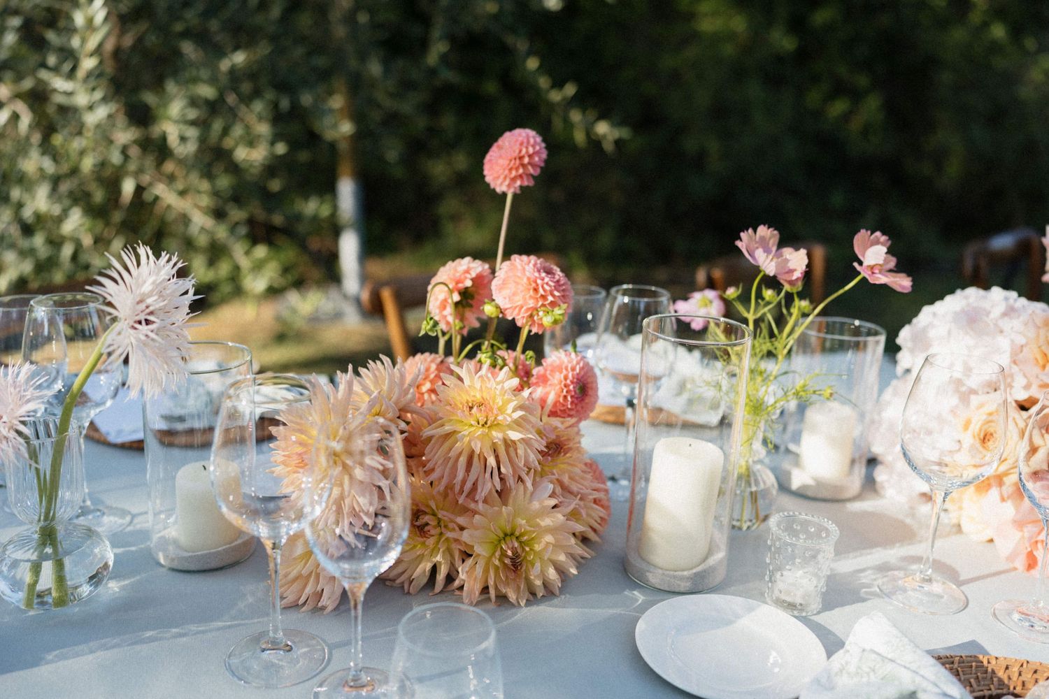 Elegant outdoor wedding table with pink dahlia centerpieces, white candles, and crystal glassware on a white linen tablecloth.