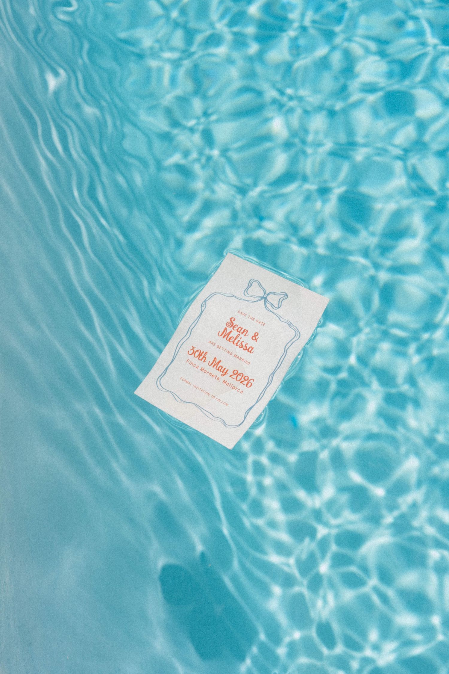 Wedding invitation card floating in a clear blue swimming pool with shimmering water reflections.