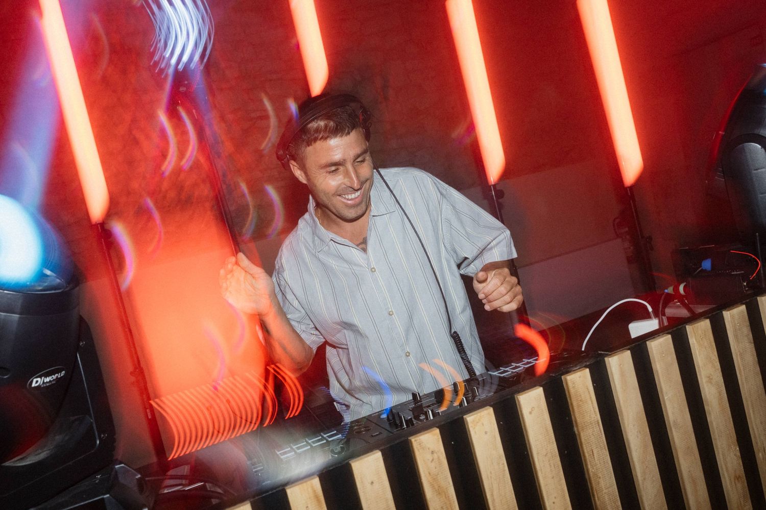 A smiling DJ performs at a nightclub behind turntables under vibrant red neon lights in a lively club setting.