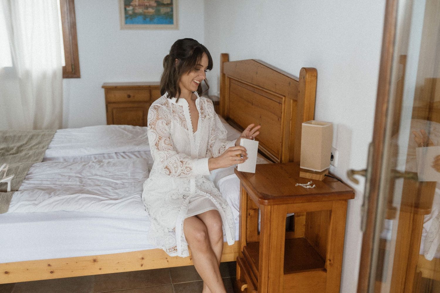 Woman in white lace robe sitting on bed reading a note by wooden nightstand in bright bedroom.