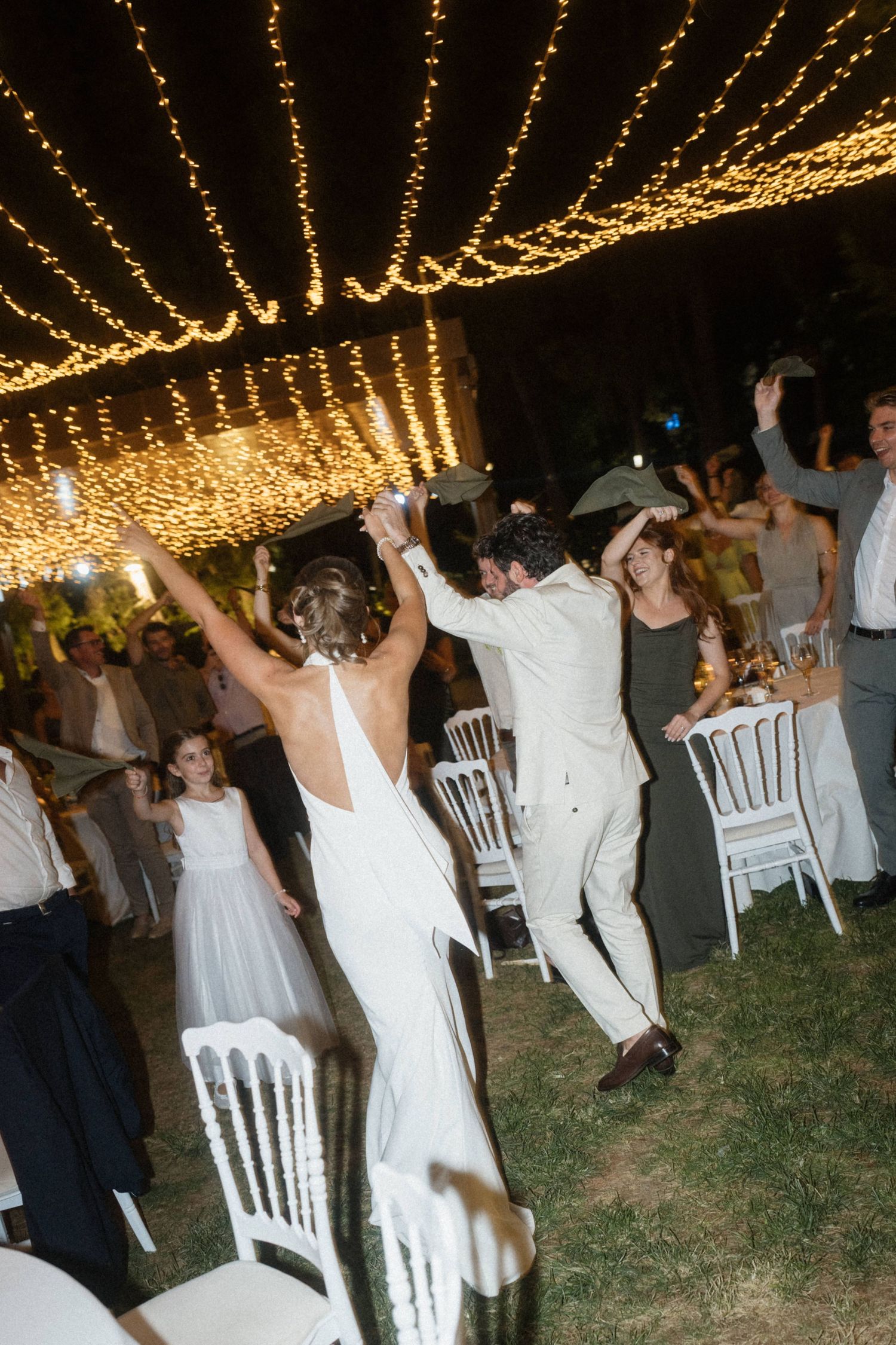Bride and groom dancing joyfully under fairy lights at an outdoor wedding reception with guests celebrating.