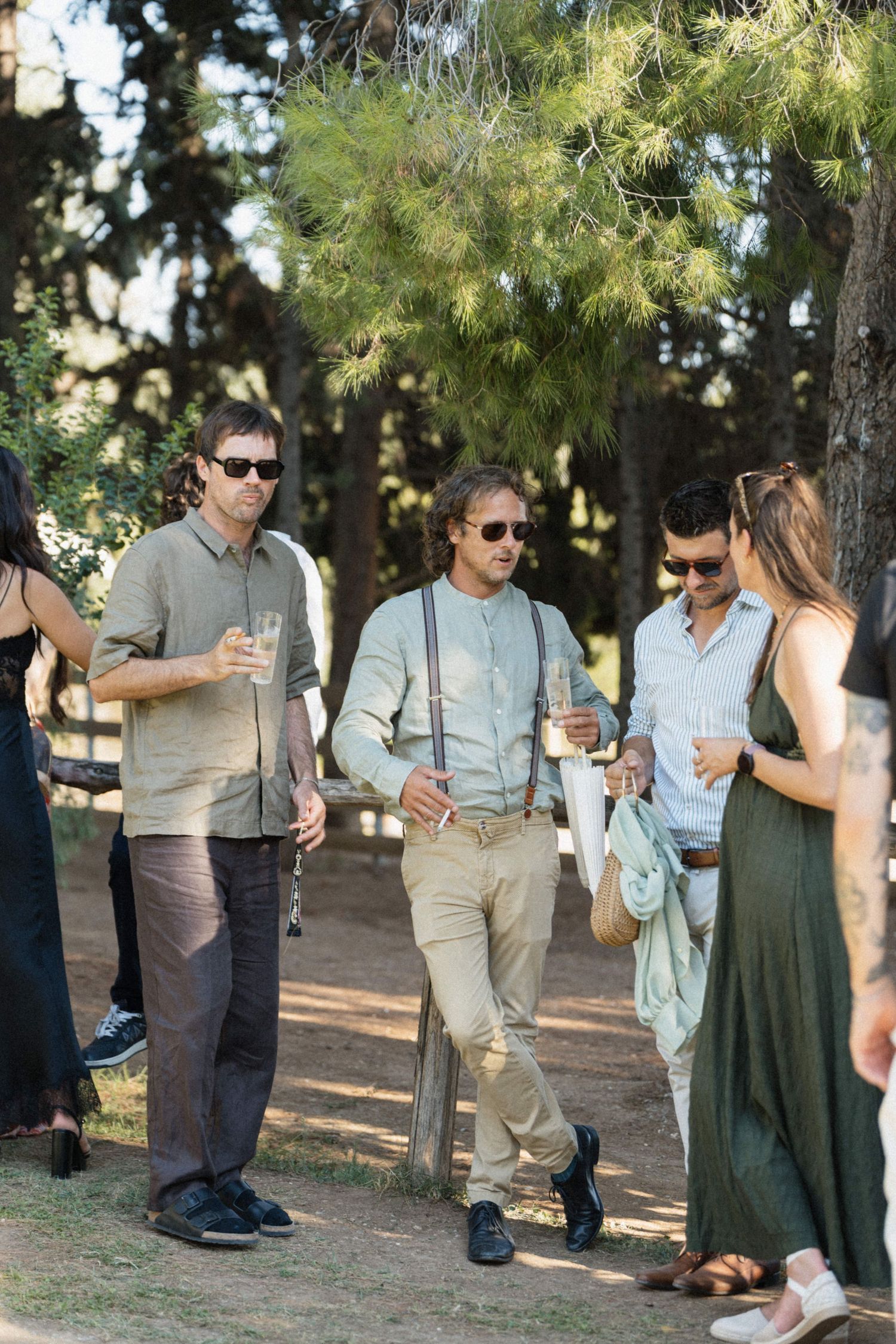 Wedding guests in smart casual attire socializing outdoors among tall pine trees at a garden reception.