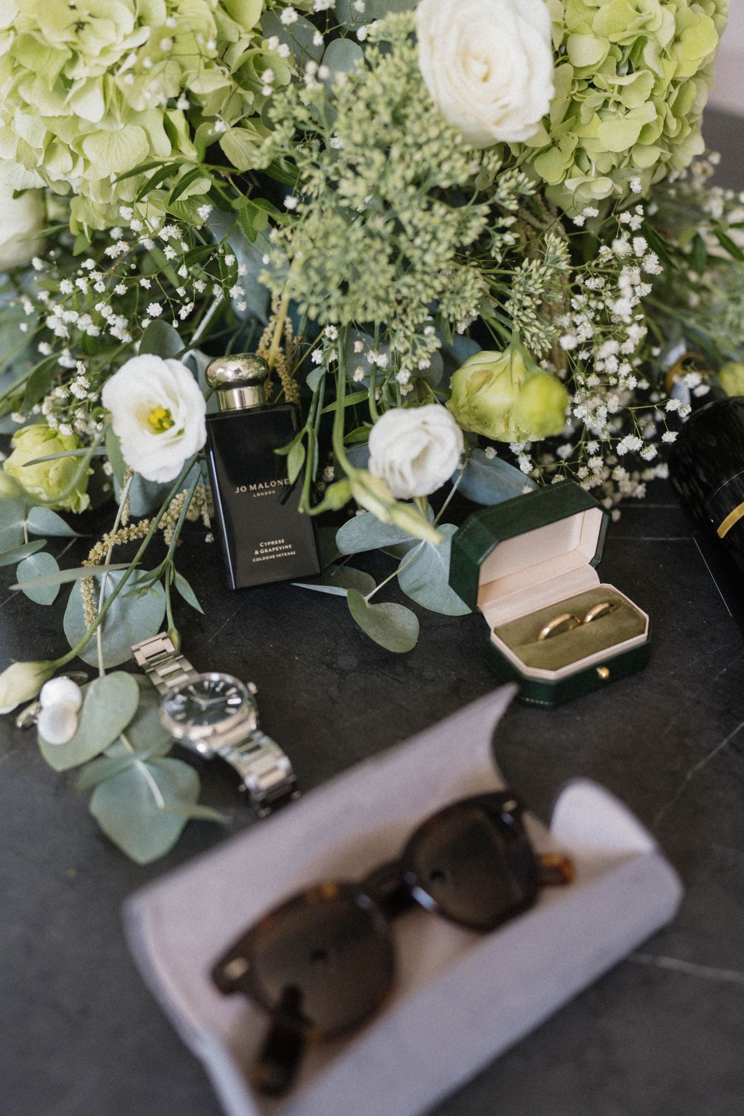 Wedding flat lay with rings, Jo Malone perfume, watch, sunglasses, and white green floral bouquet on dark surface.