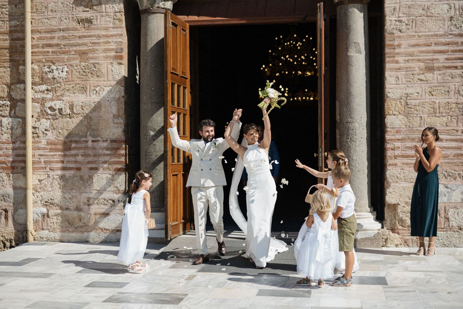 Joyful newlyweds exit a historic church as guests toss confetti, with flower girls in white dresses celebrating nearby.