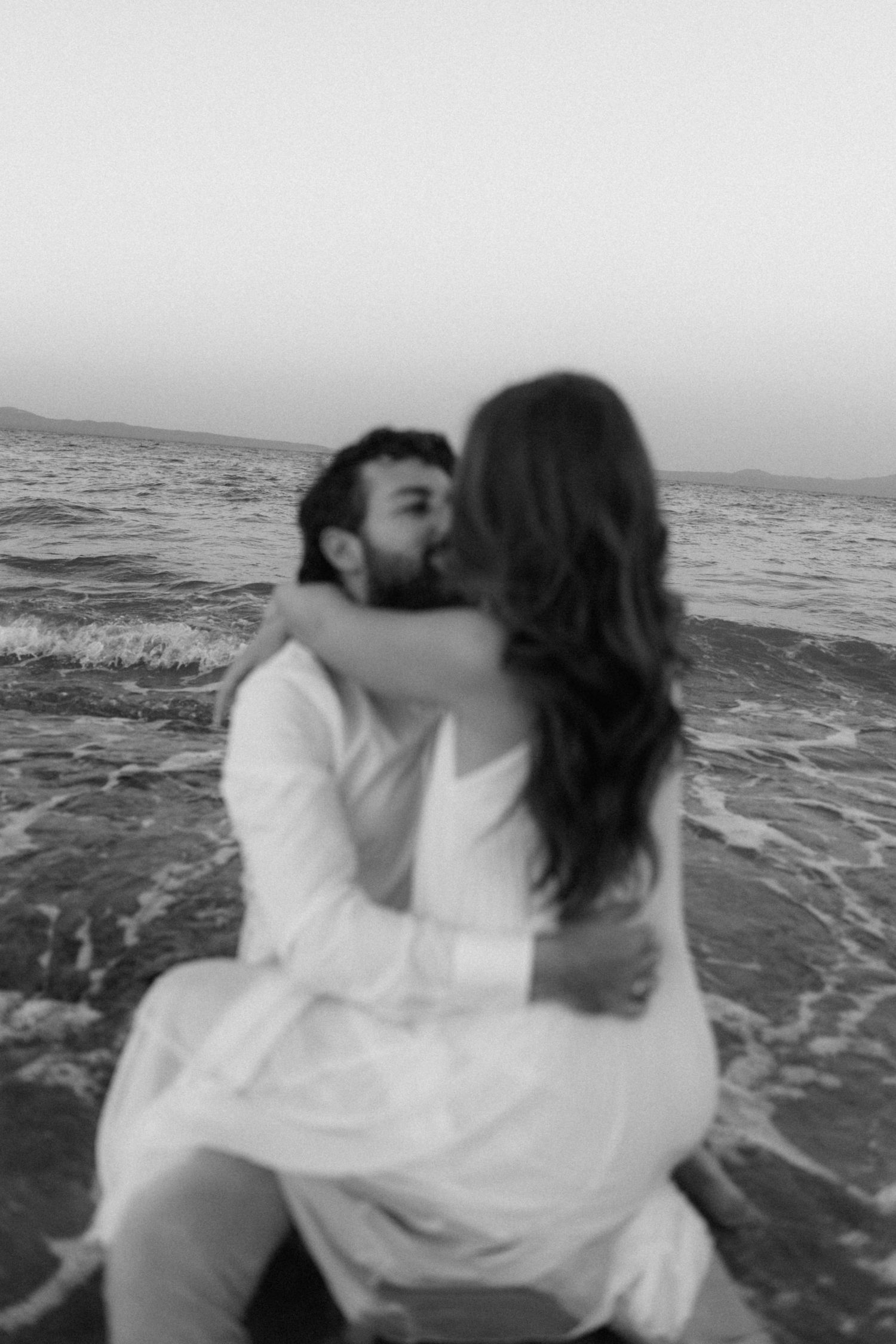 Black and white photo of couple kissing while sitting in ocean waves on a beach at dusk.