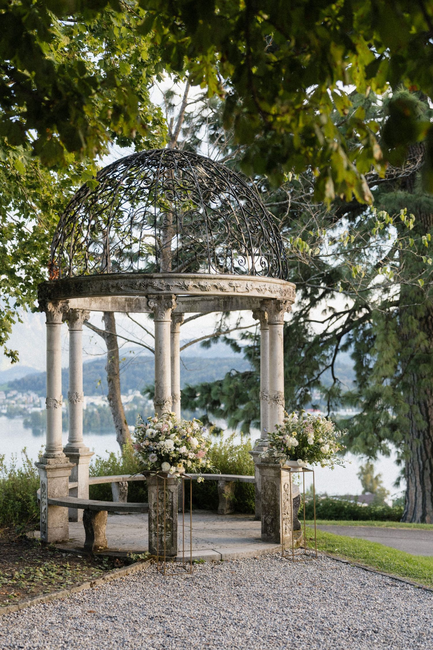 Ornate stone gazebo adorned with white floral arrangements overlooking a serene lake surrounded by lush trees.