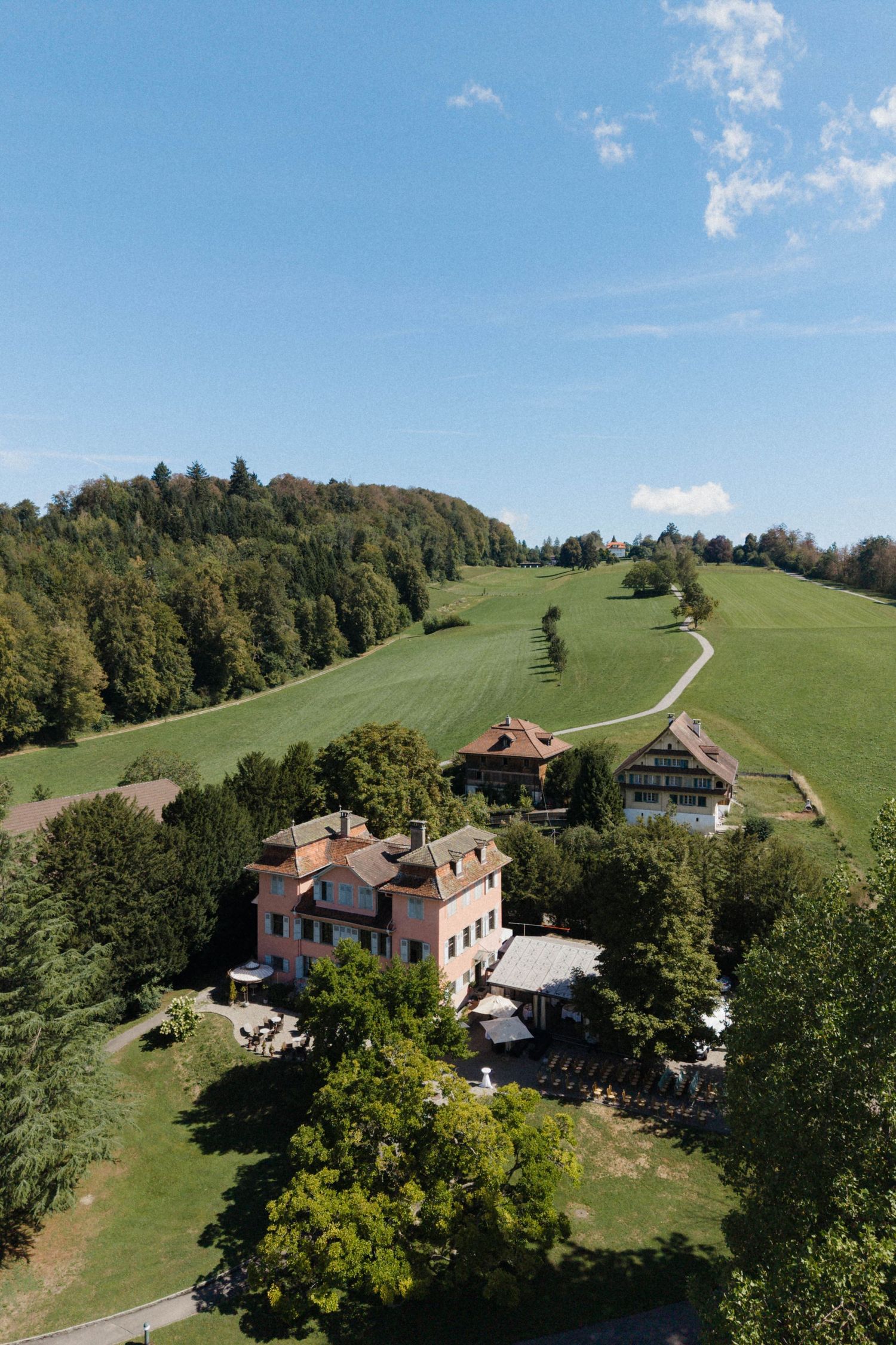 Aerial view of a charming countryside estate surrounded by lush green hills and dense forest under a clear blue sky.