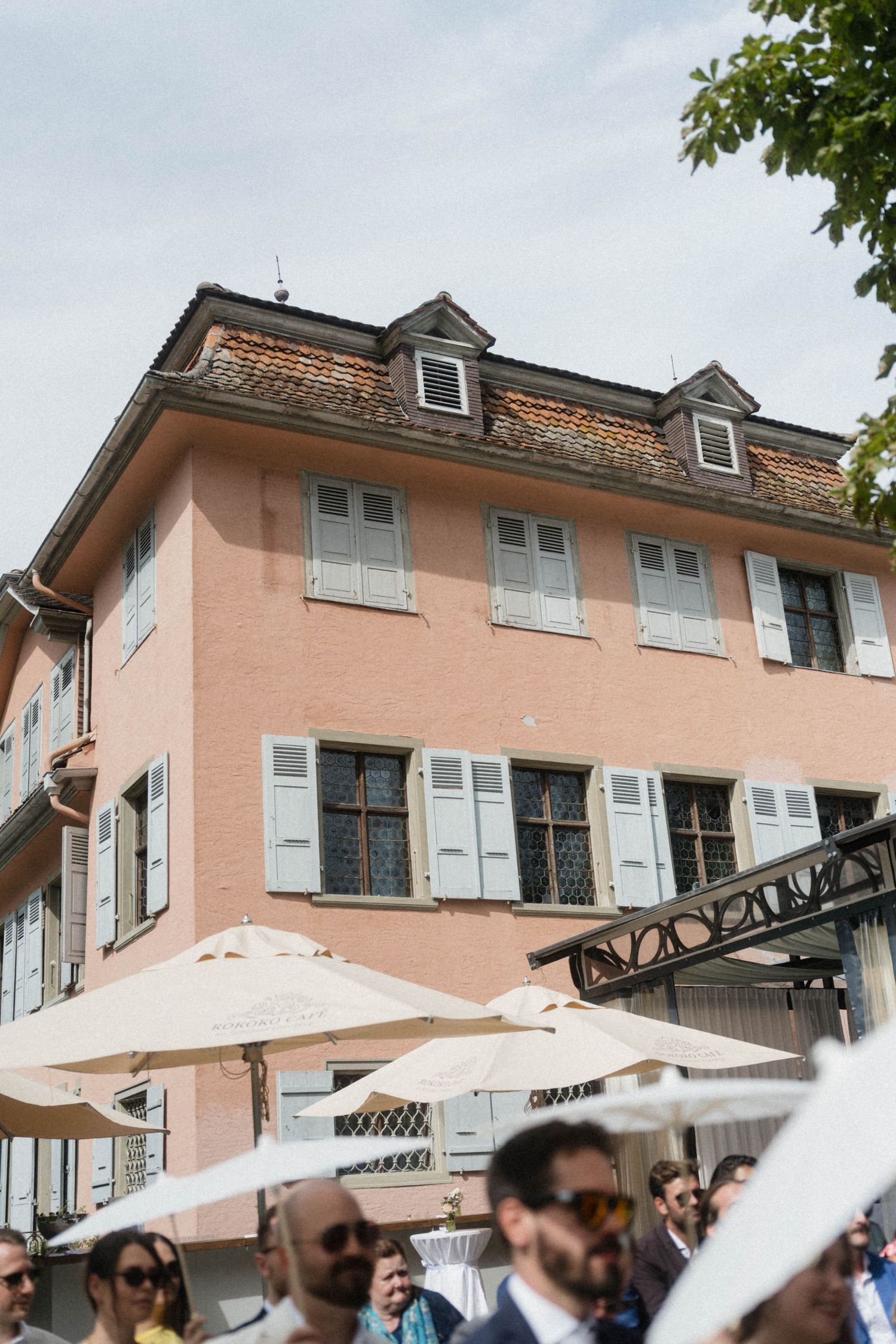 Pink European building with dormer windows and white shutters, outdoor dining umbrellas and wedding guests gathered below.