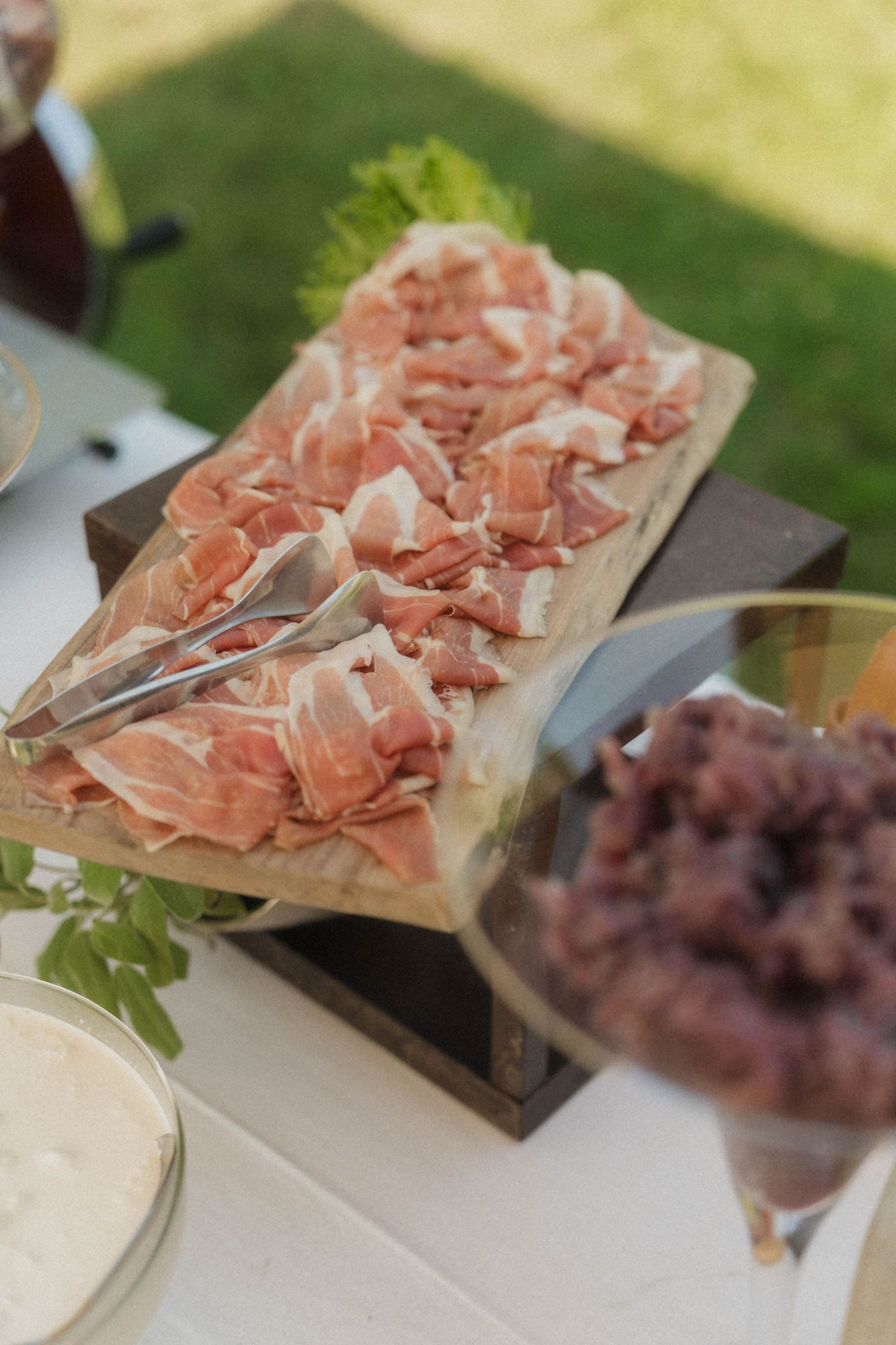 Sliced prosciutto displayed on wooden board with tongs at outdoor wedding reception catering table.