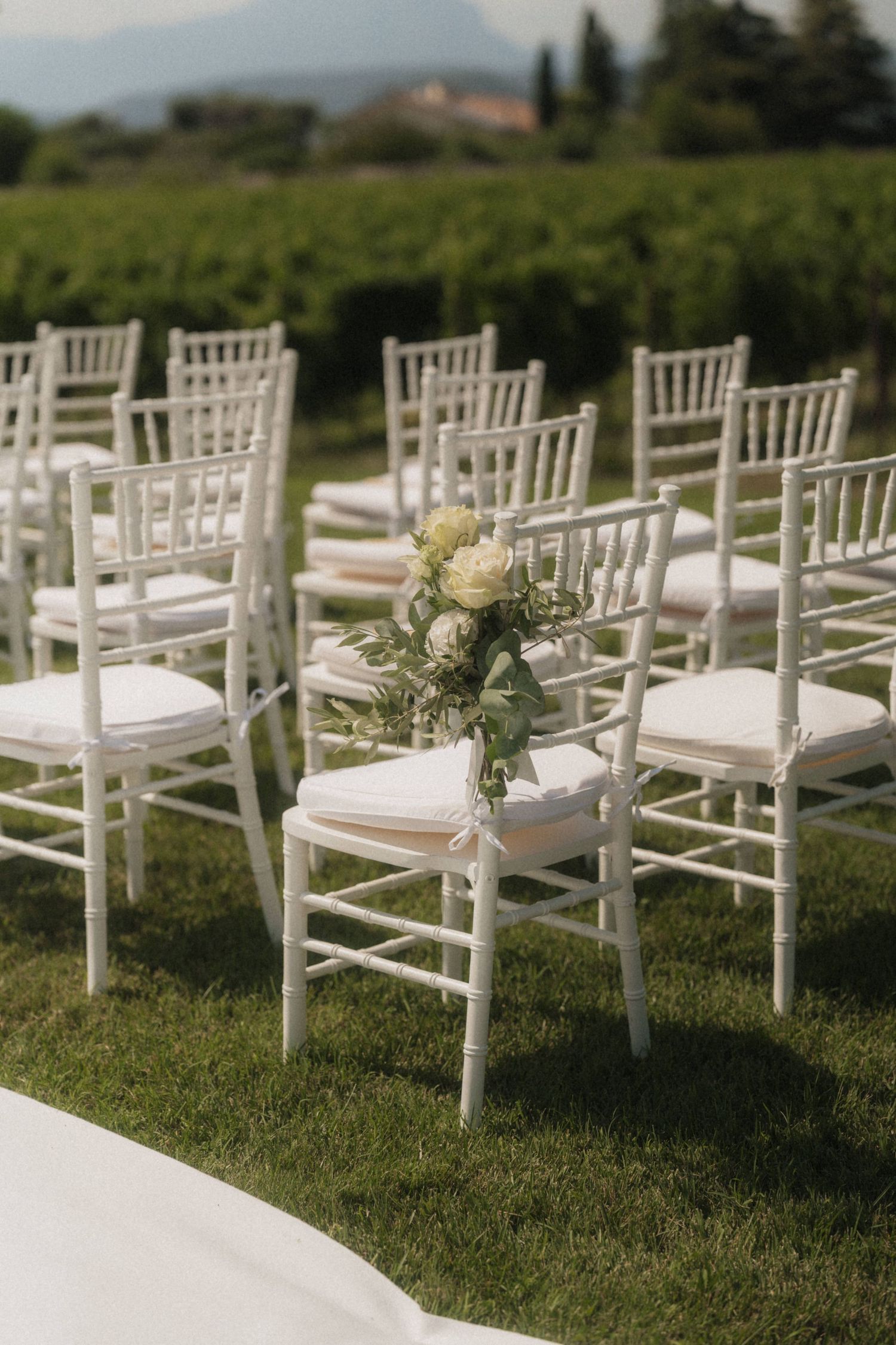 White Chiavari chairs with floral arrangements set up on green lawn for an elegant outdoor wedding ceremony.
