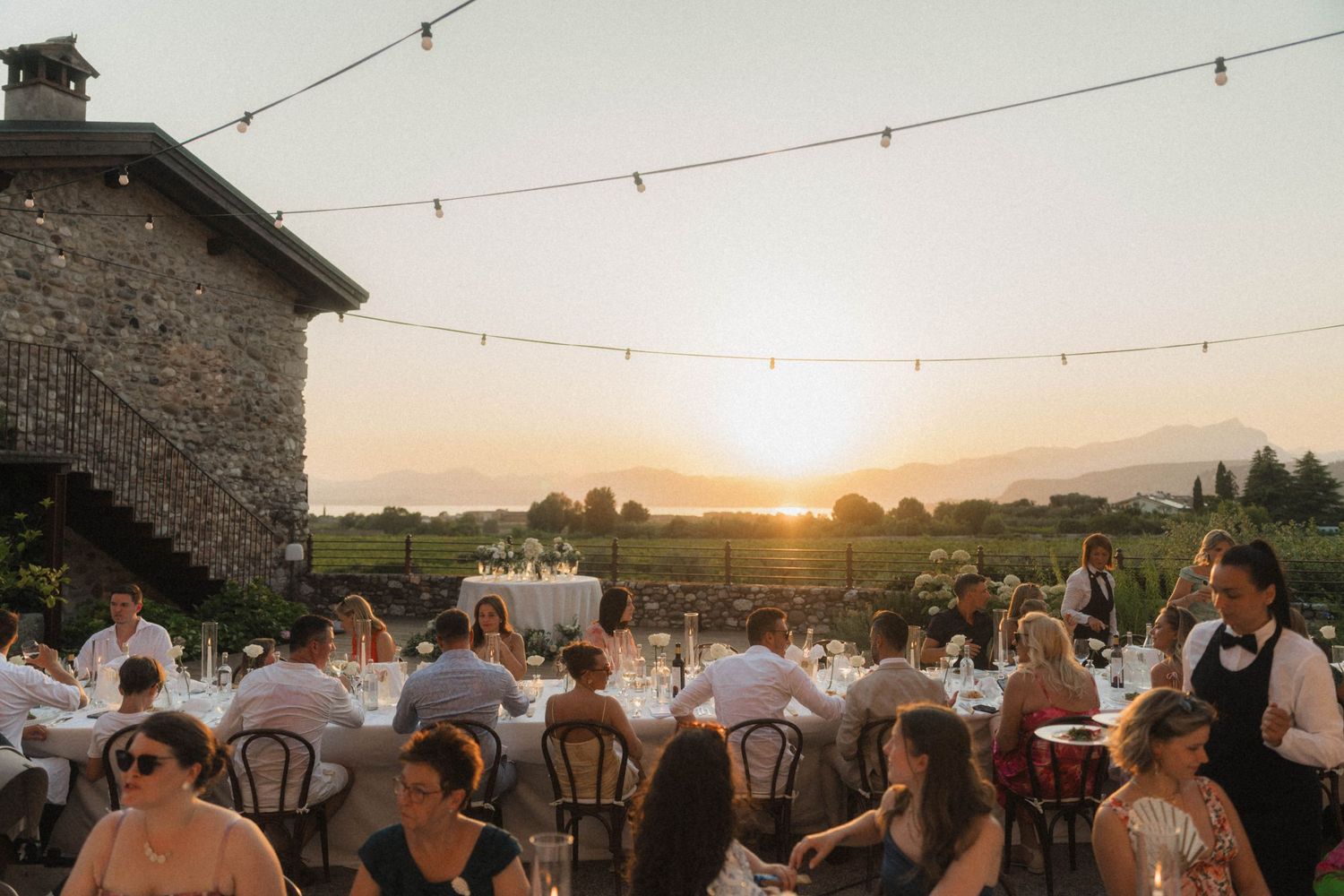 Wedding guests dining outdoors at sunset with string lights and mountain views at a rustic vineyard venue.