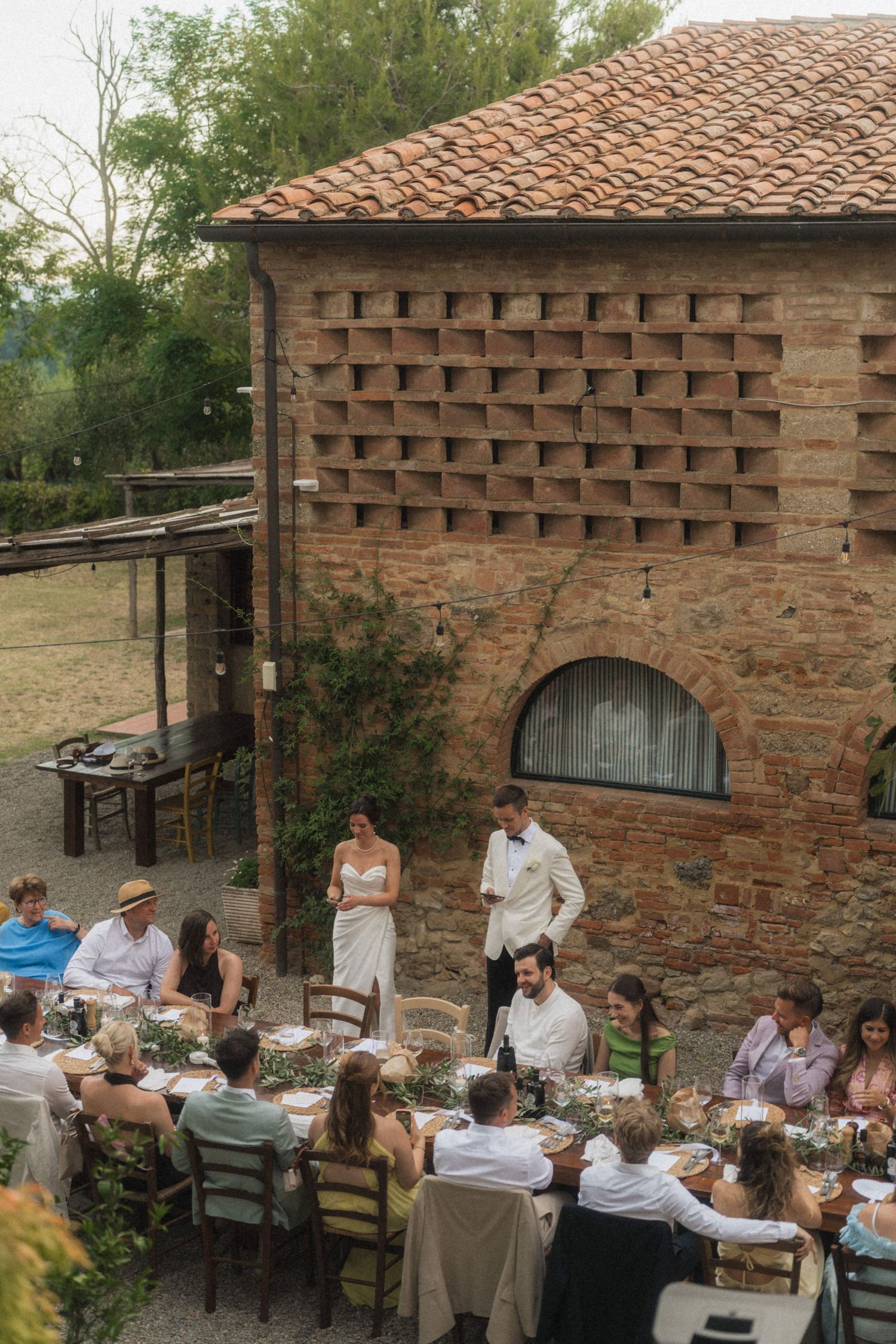 Aerial view of wedding guests dining outdoors beside rustic Tuscan brick farmhouse on sunny day.