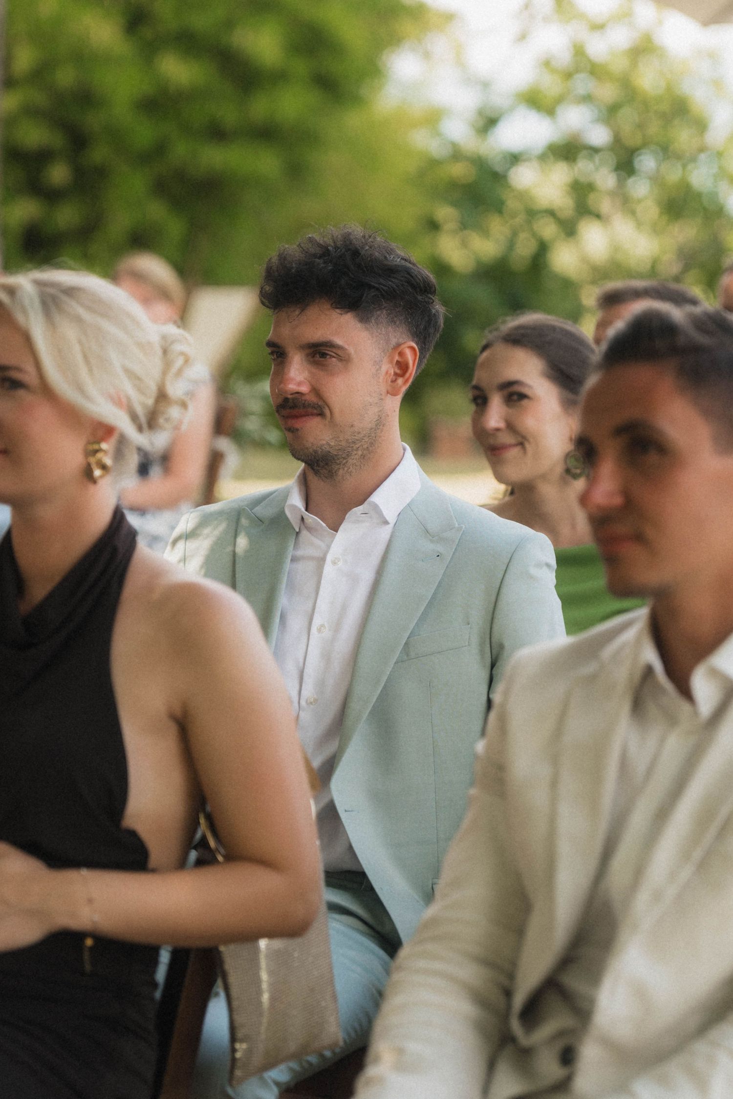 Wedding guests seated outdoors, man in light blue suit smiling during garden ceremony.