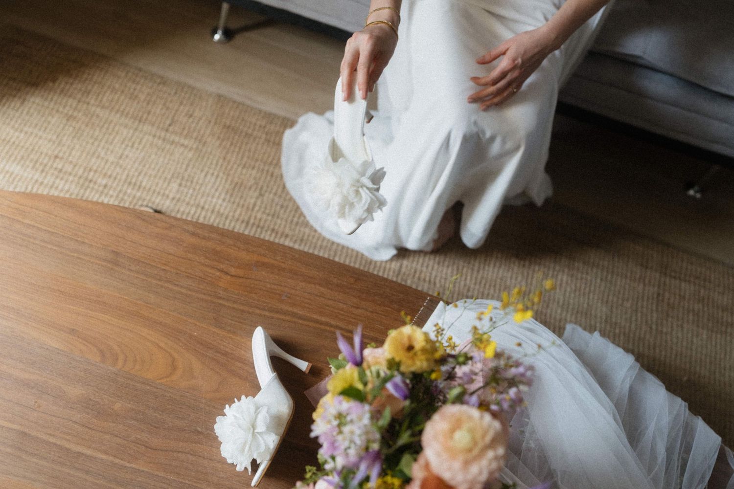 Bride in white wedding dress picking up bridal shoes near a wooden table with colorful flower bouquet.