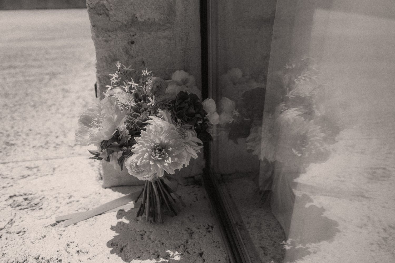 Black and white photo of a floral bouquet with daisies and mixed flowers resting against a stone wall and glass window.