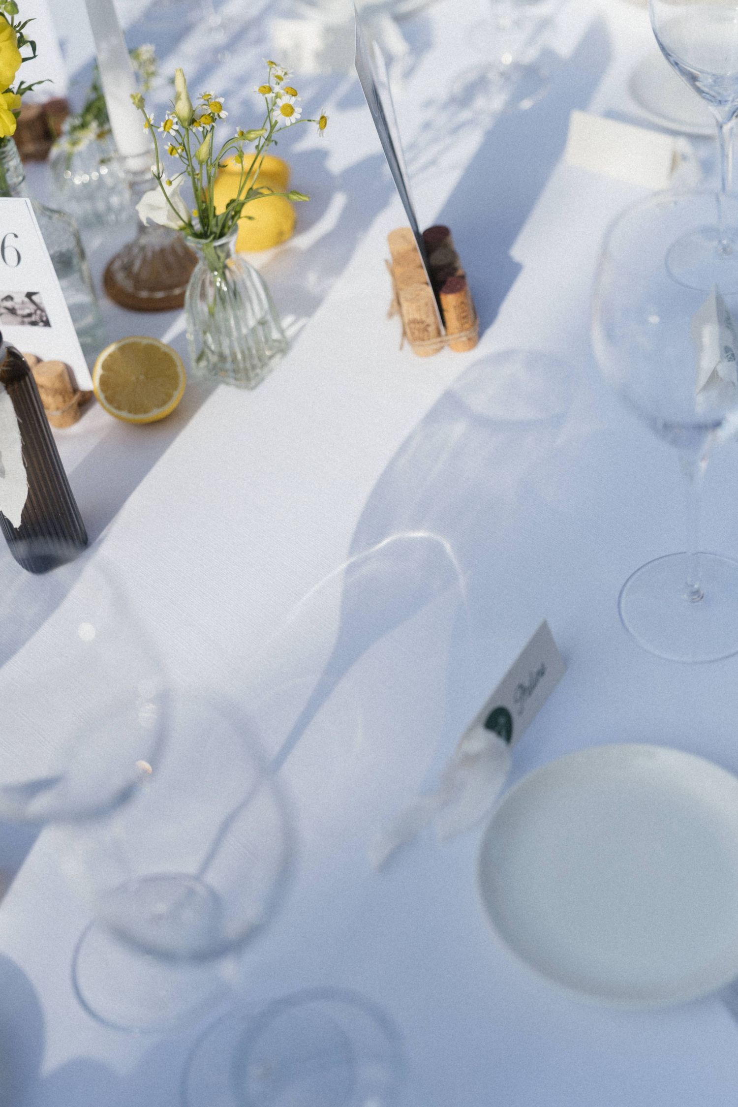 Elegant white wedding table setting with wine glasses, lemons, wildflowers in vases, and place cards in bright sunlight.