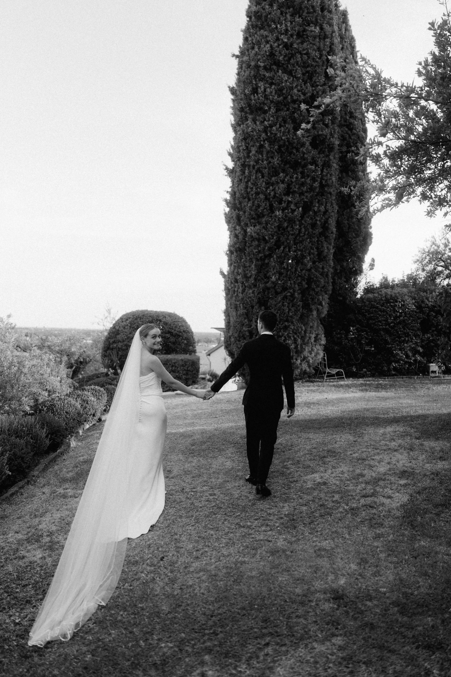 Black and white photo of bride and groom holding hands walking toward a tall cypress tree in an Italian garden.