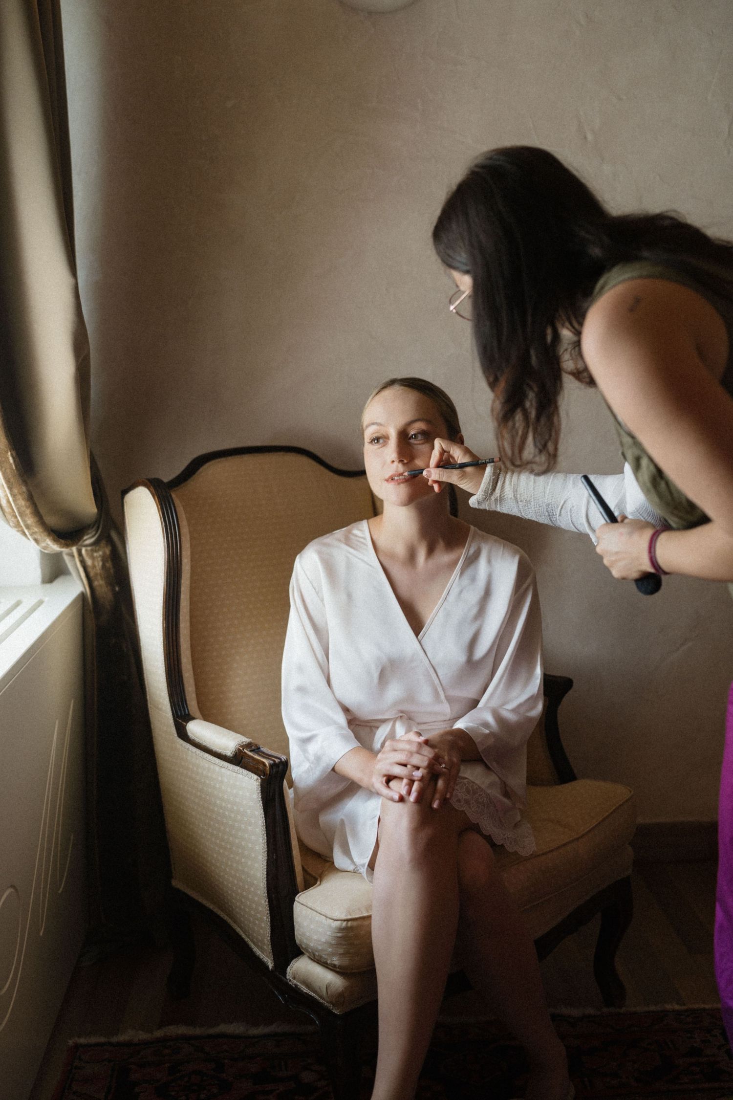 A makeup artist applies makeup to a bride in a white robe seated in an elegant chair by a window.