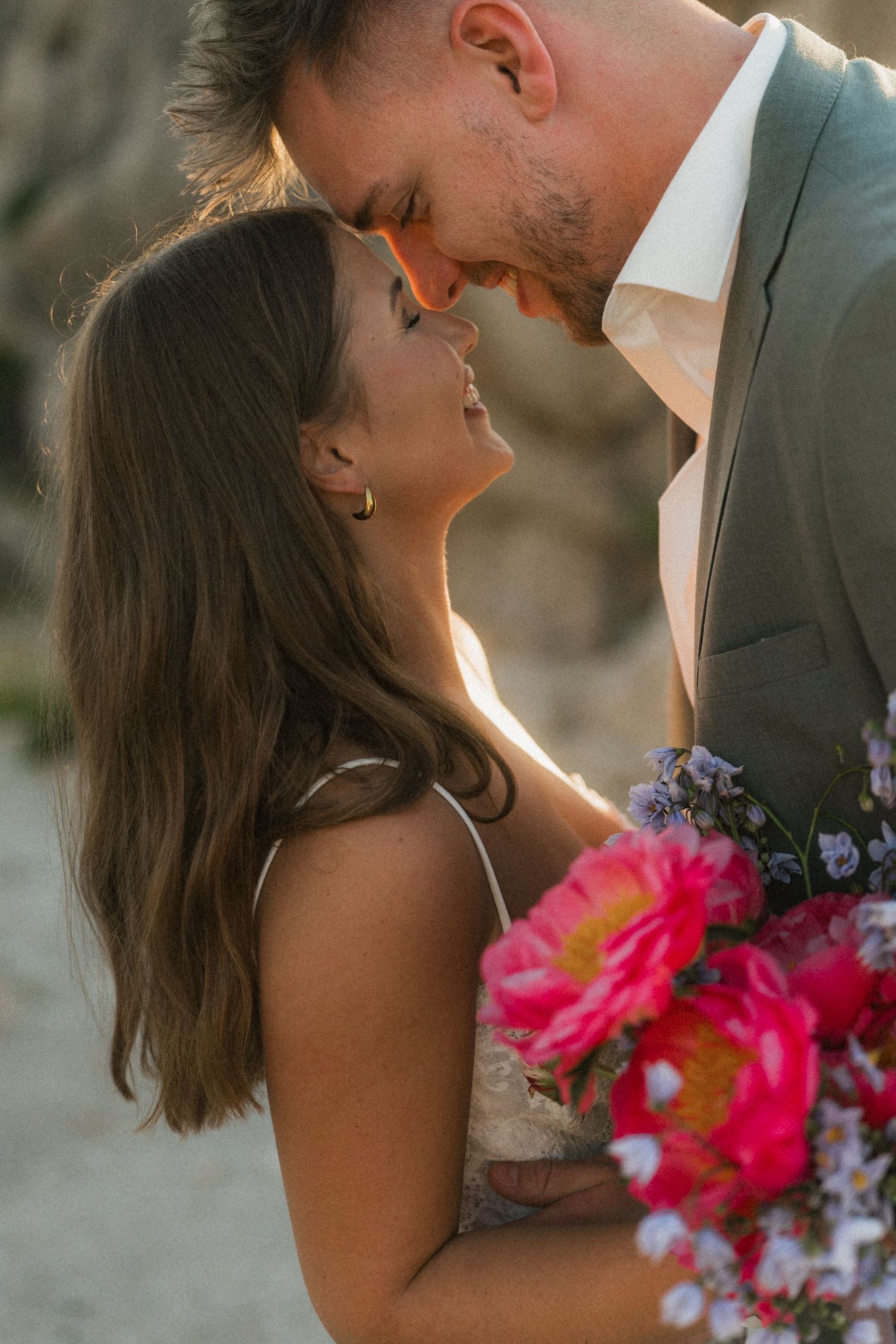 Romantic close-up of bride and groom touching foreheads at sunset, bride holding vibrant pink peony wedding bouquet.