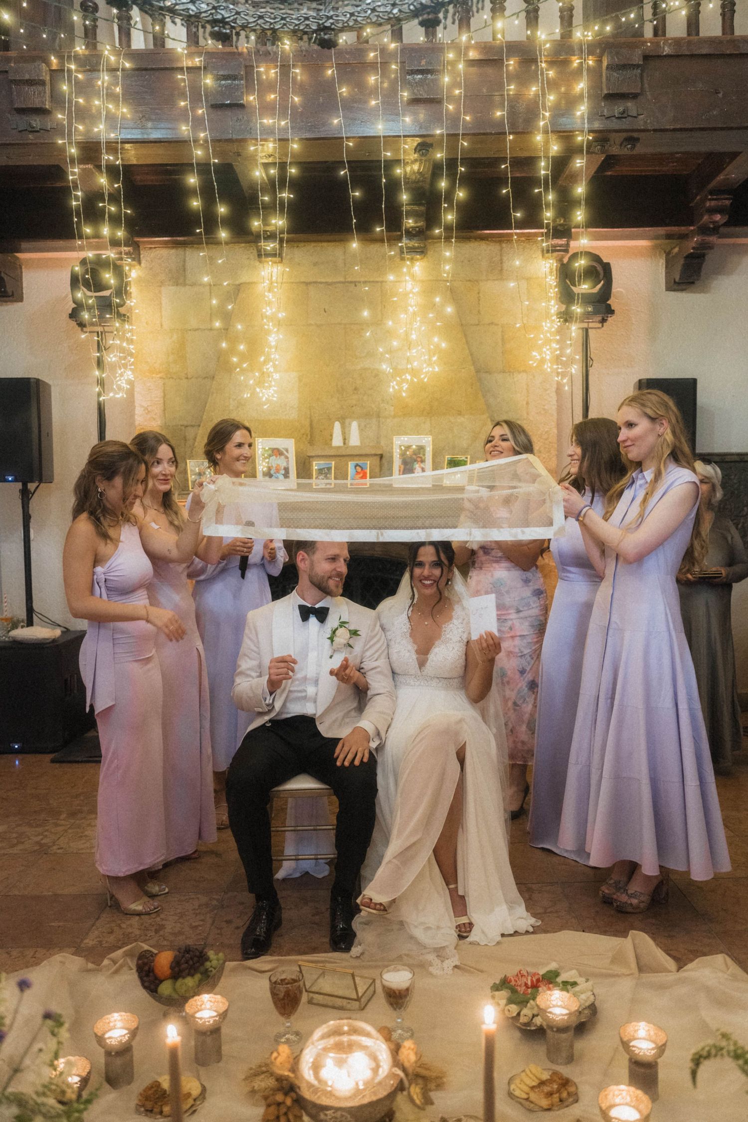 Bride and groom seated as bridesmaids in lavender dresses hold veil under fairy lights.