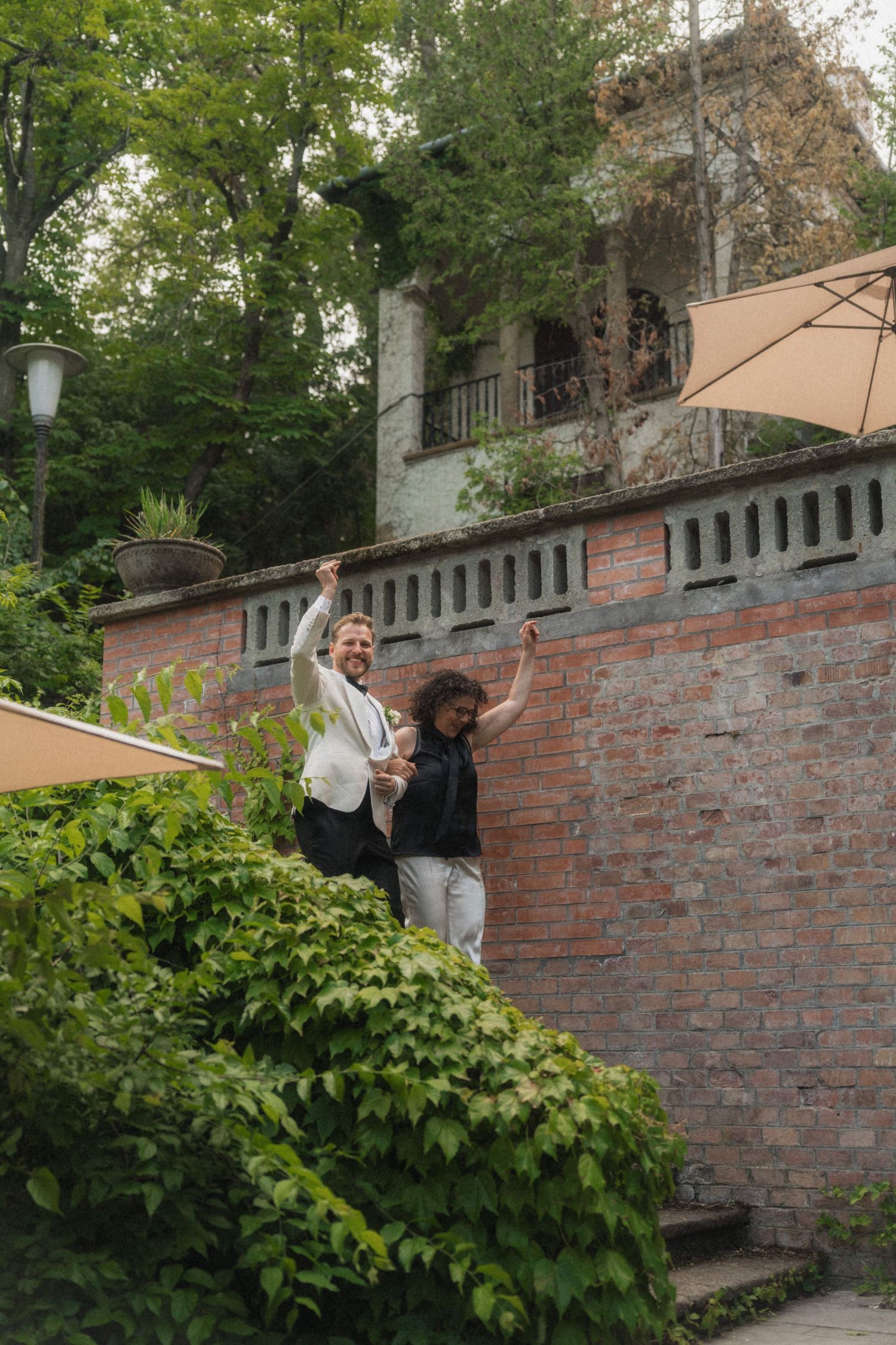 Two joyful people raising their arms outside a brick wall surrounded by lush greenery.