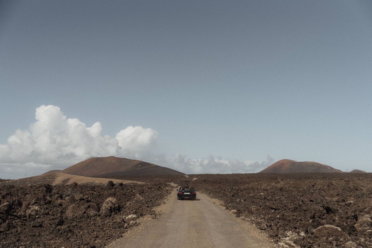 A lone vehicle drives along a dirt road through a stark volcanic landscape with rocky terrain and hills under a partly cloudy sky.