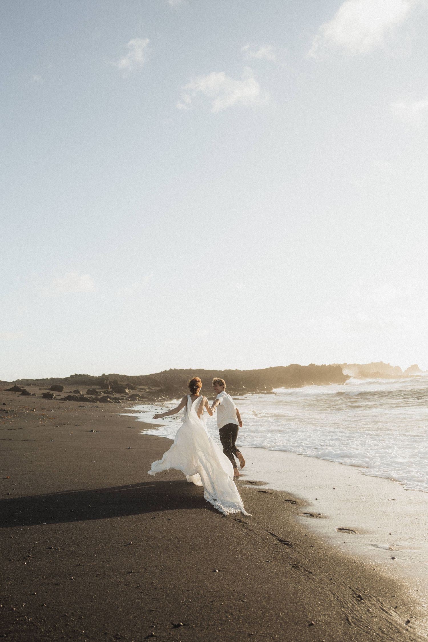 Bride and groom walk hand in hand along black sand beach at sunset.