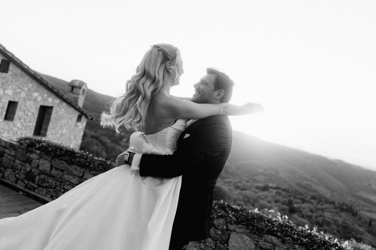 Black and white romantic photo of a couple embracing near a stone building with rolling hills at sunset.