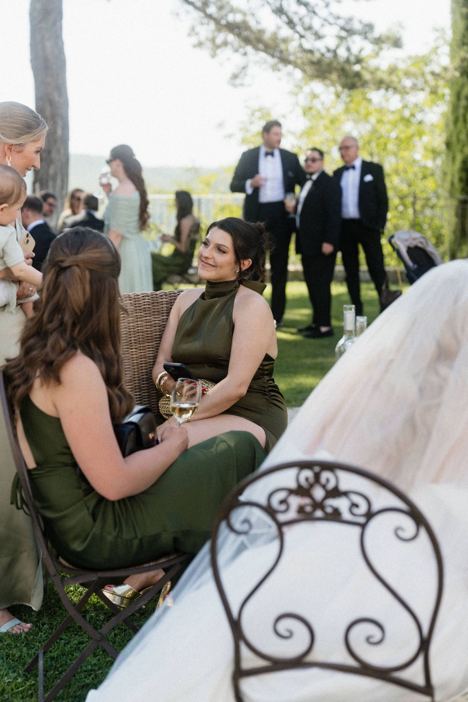 Wedding guests in olive green dresses socializing and enjoying champagne at an outdoor garden reception.