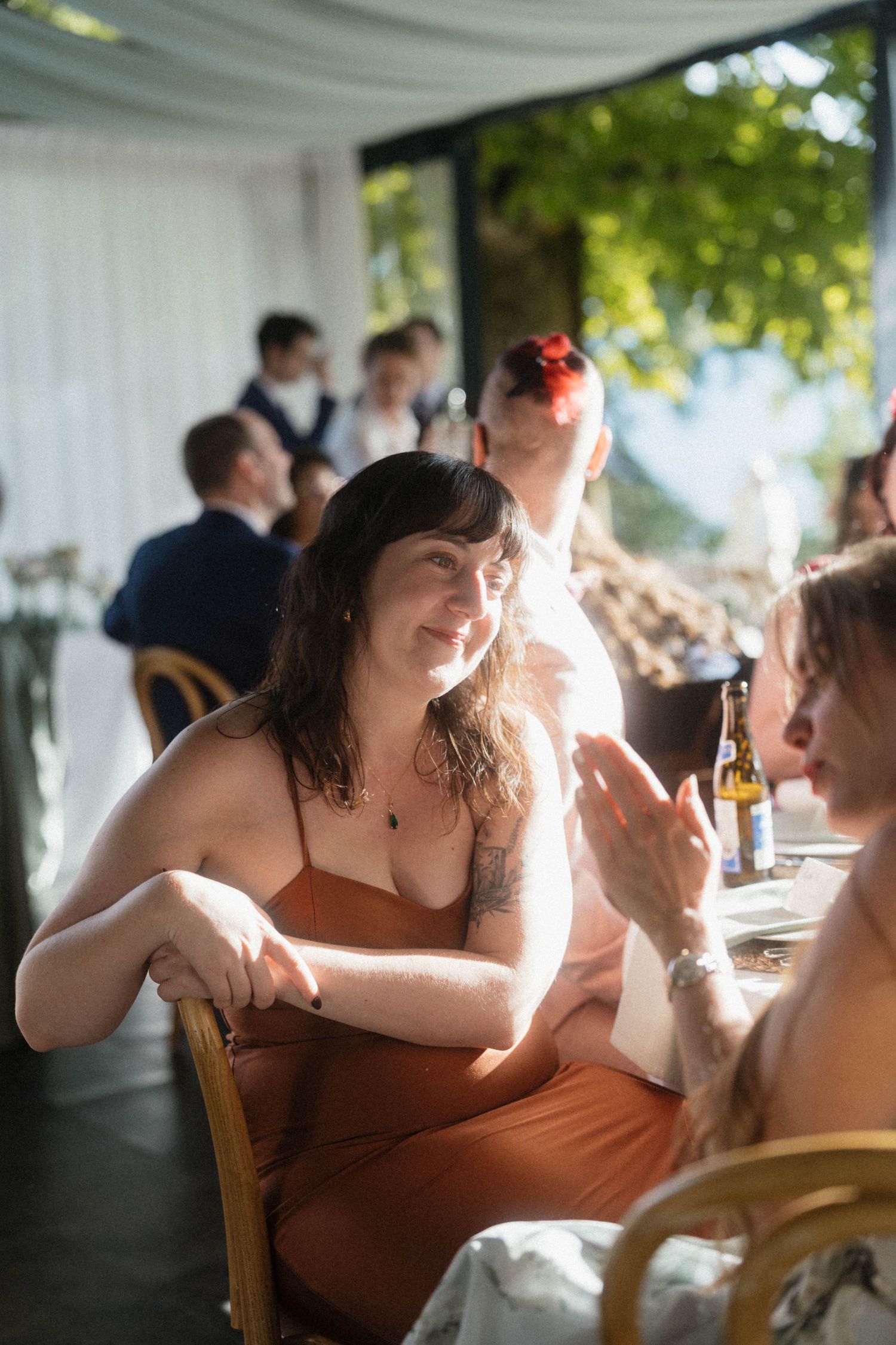 Woman in rust orange dress smiling and conversing at a sunlit outdoor wedding reception dinner table.
