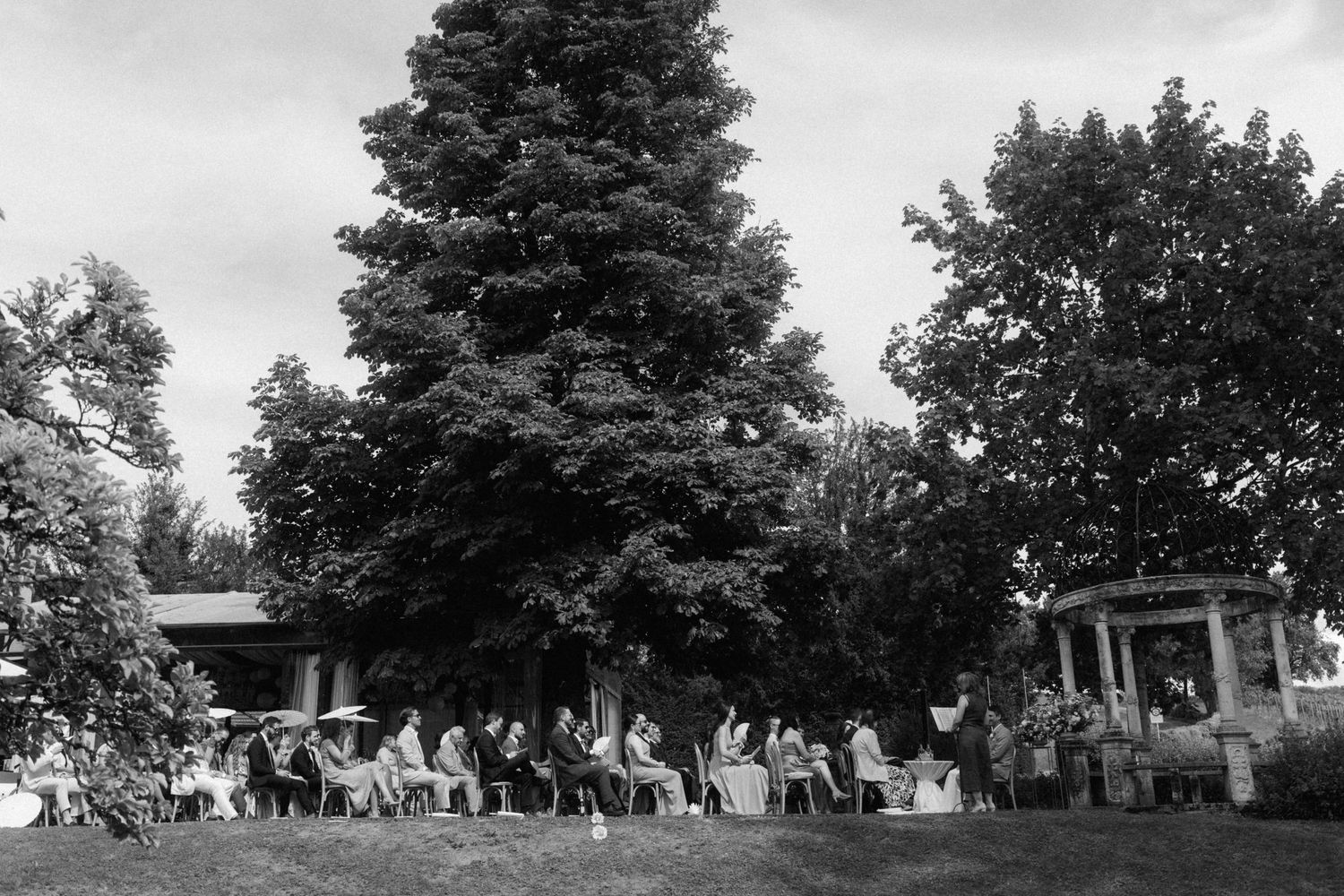 Black and white wide shot of wedding guests seated outdoors beneath large trees for an elegant garden ceremony.