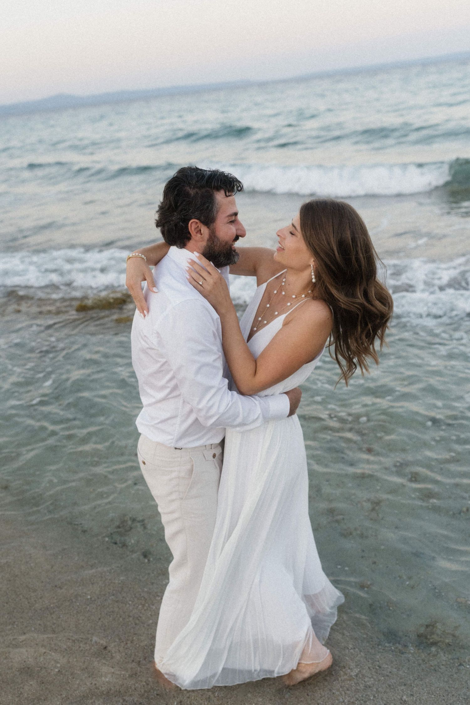 Smiling couple in white wedding attire embrace romantically on beach with ocean waves crashing behind them.
