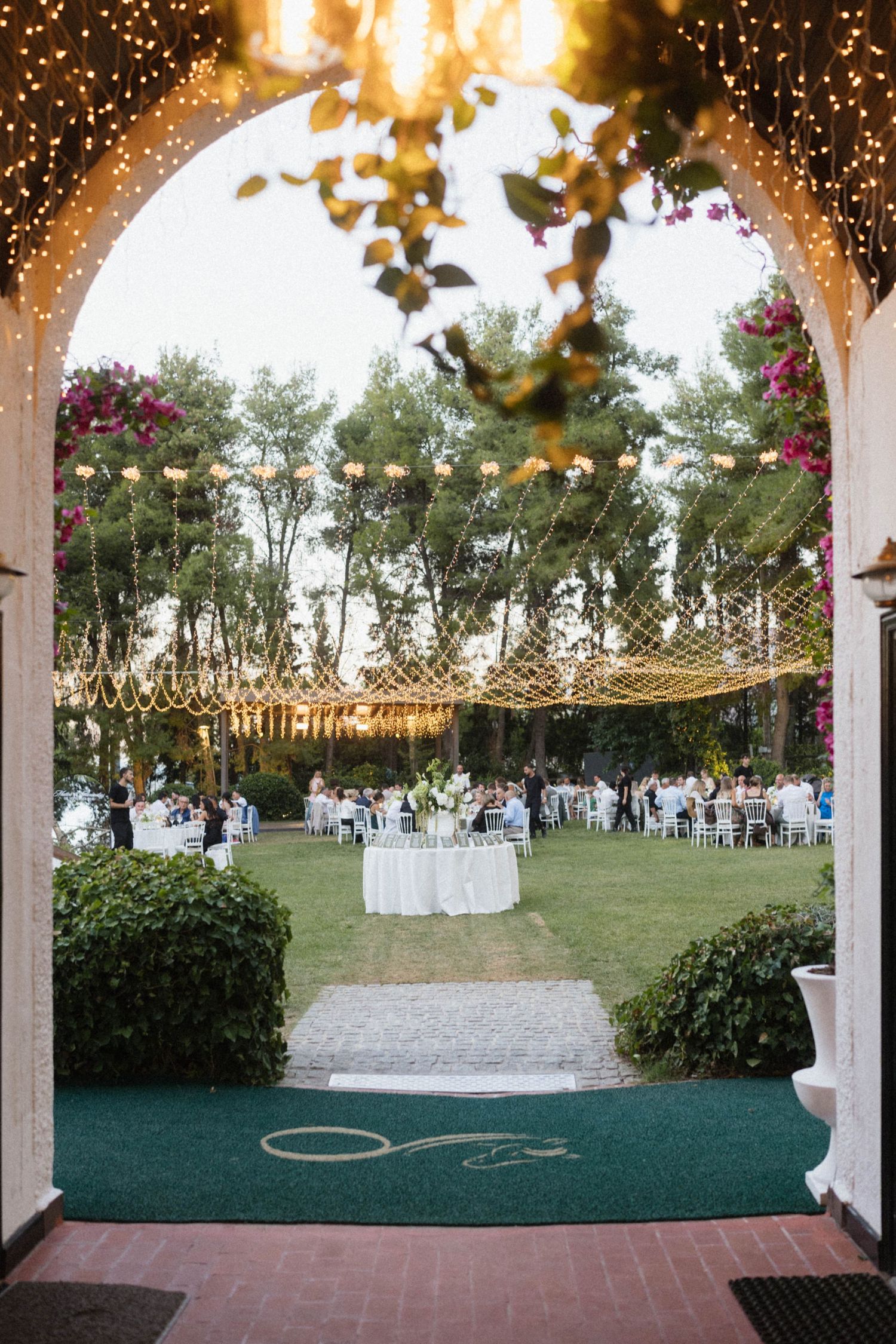 Romantic outdoor wedding reception viewed through arched entrance with string lights and white round tables on green lawn.