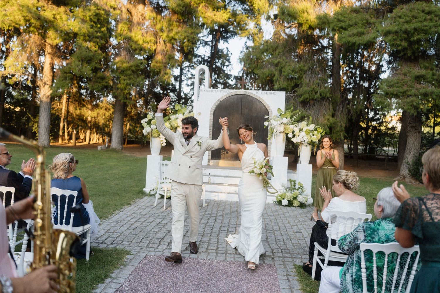 Newlyweds celebrate joyfully walking down aisle at outdoor garden wedding with white floral decorations.
