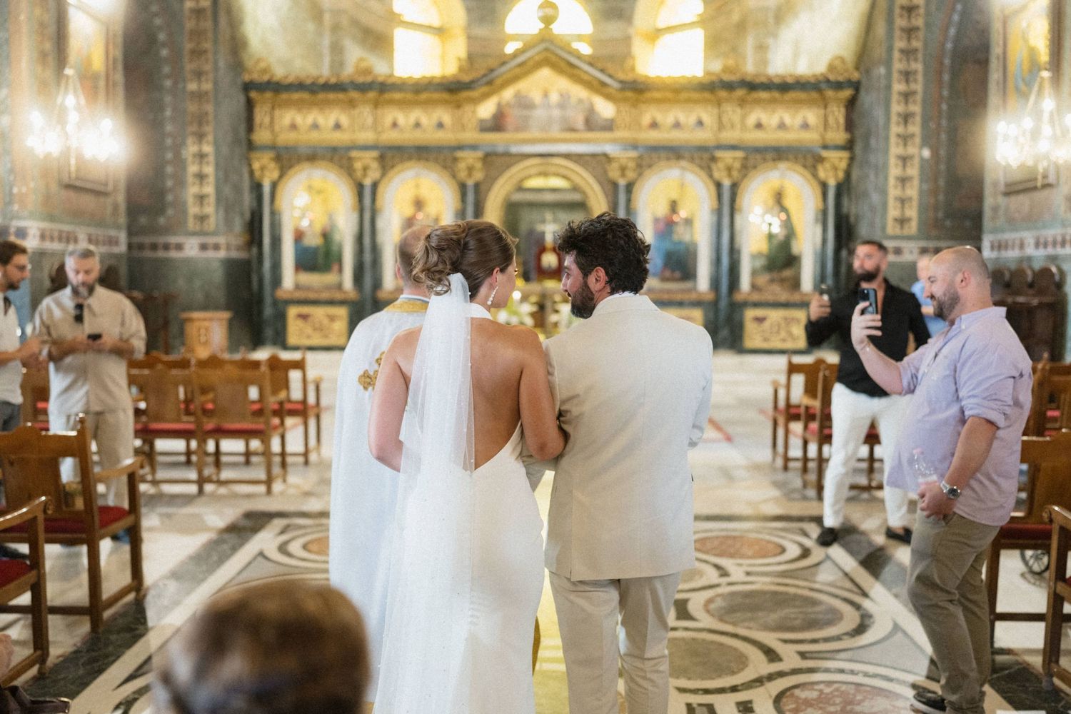 A bride and groom stand at the altar during a wedding ceremony inside an ornate Orthodox church with golden icons.