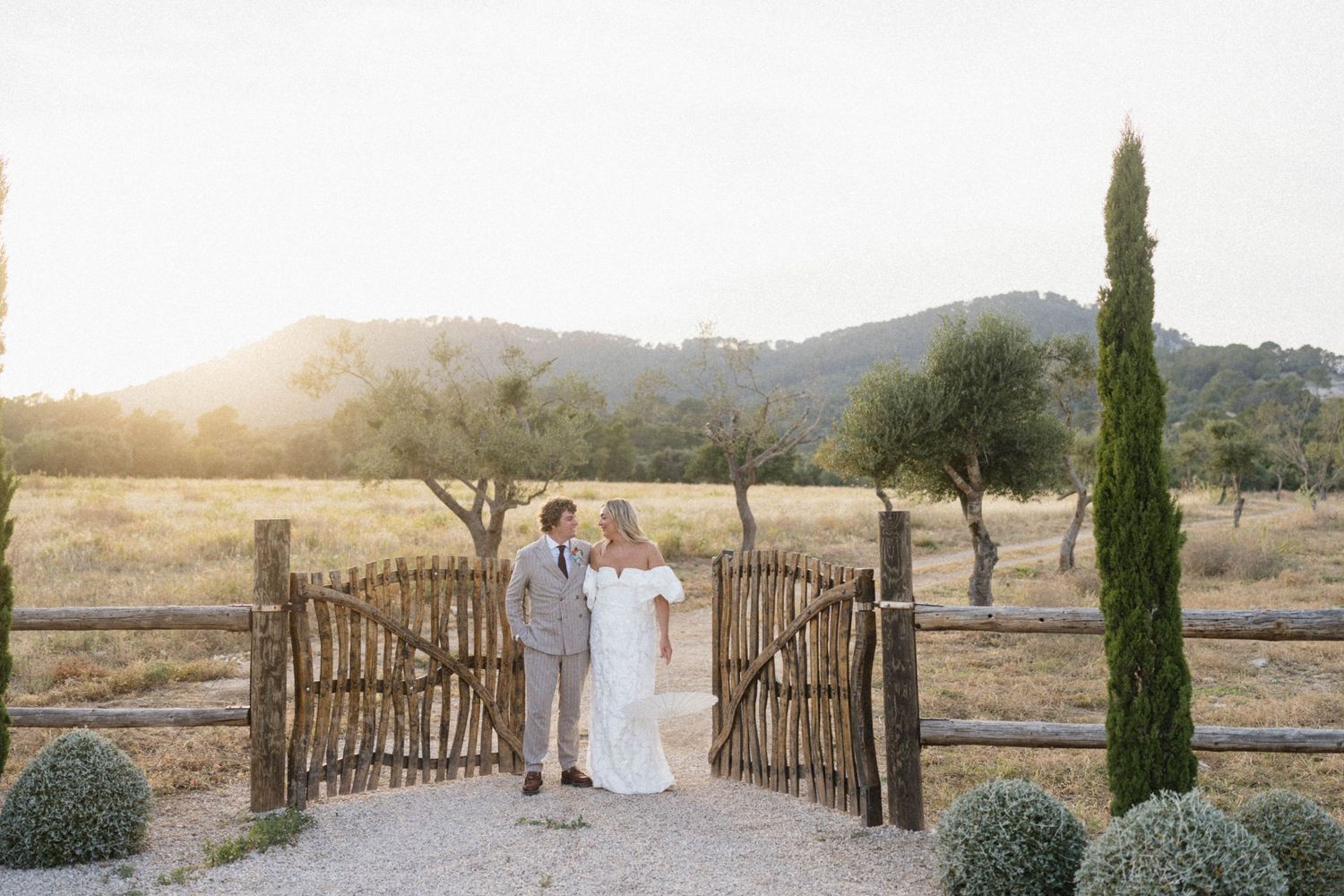 Couple poses at rustic wooden gate during golden hour with cypress trees and rolling hills at outdoor wedding venue.