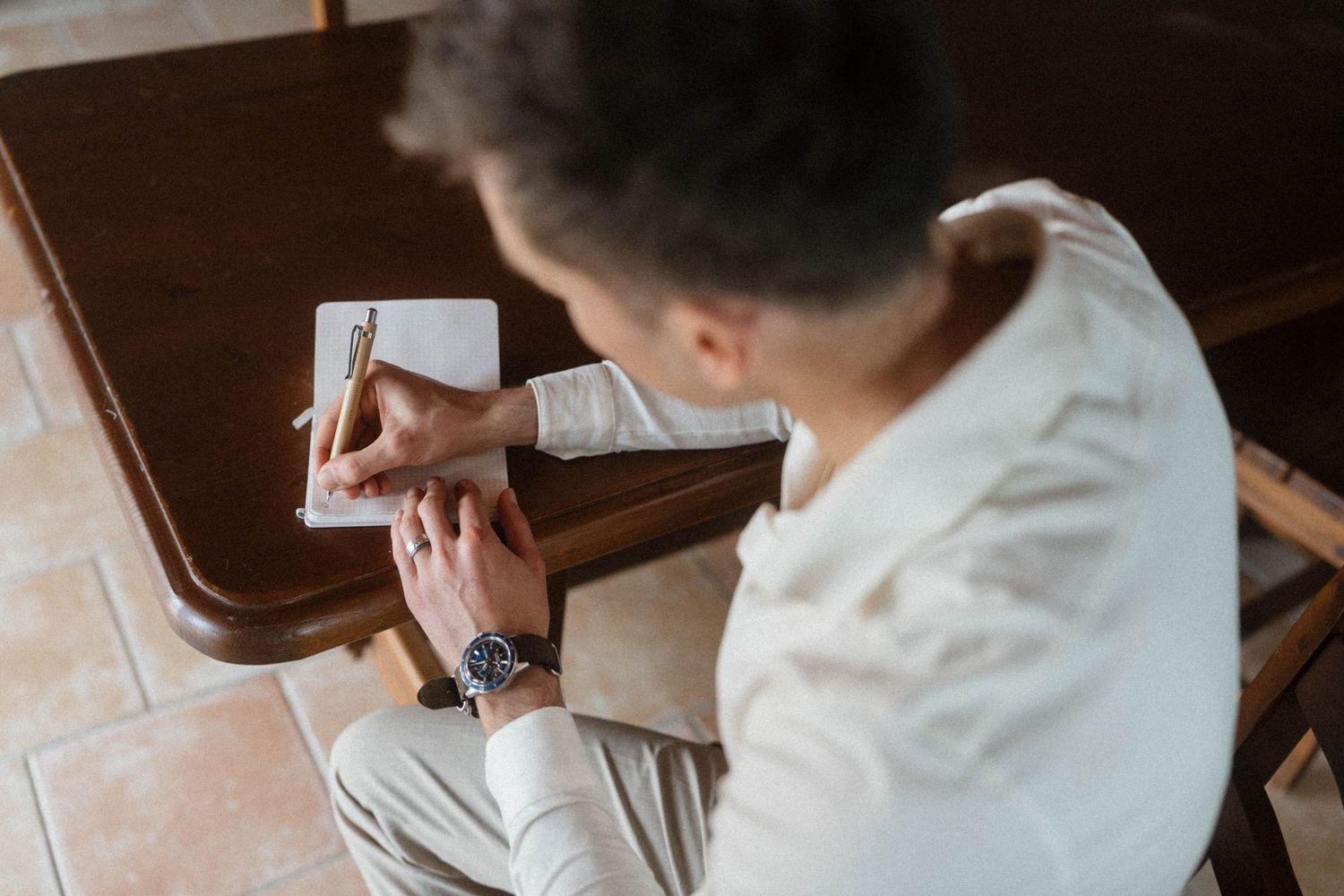 Man in white shirt writing in notebook at wooden desk, wearing a watch.