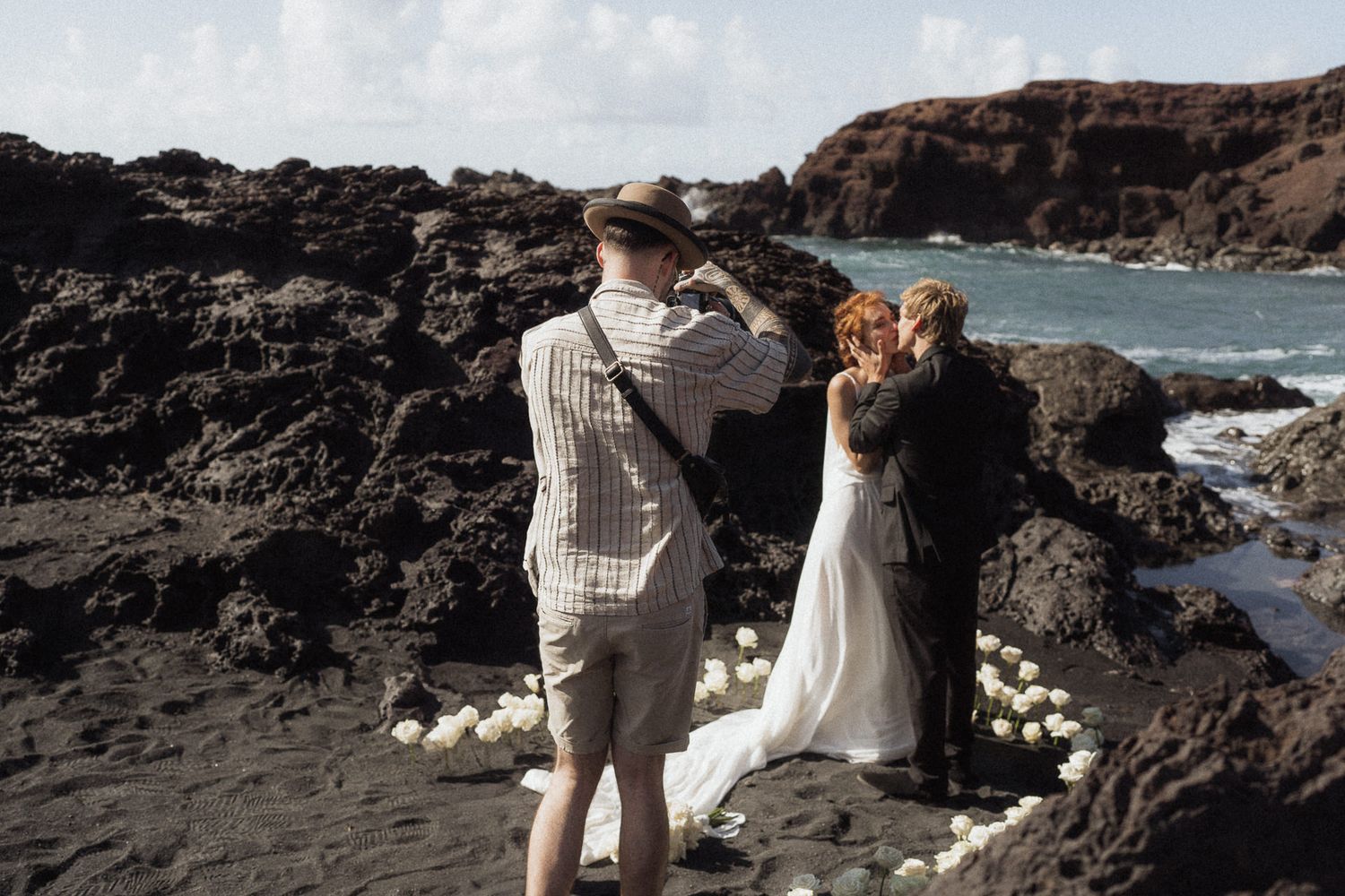 Couple sharing a kiss during a beach wedding ceremony on black volcanic rocks by the ocean.